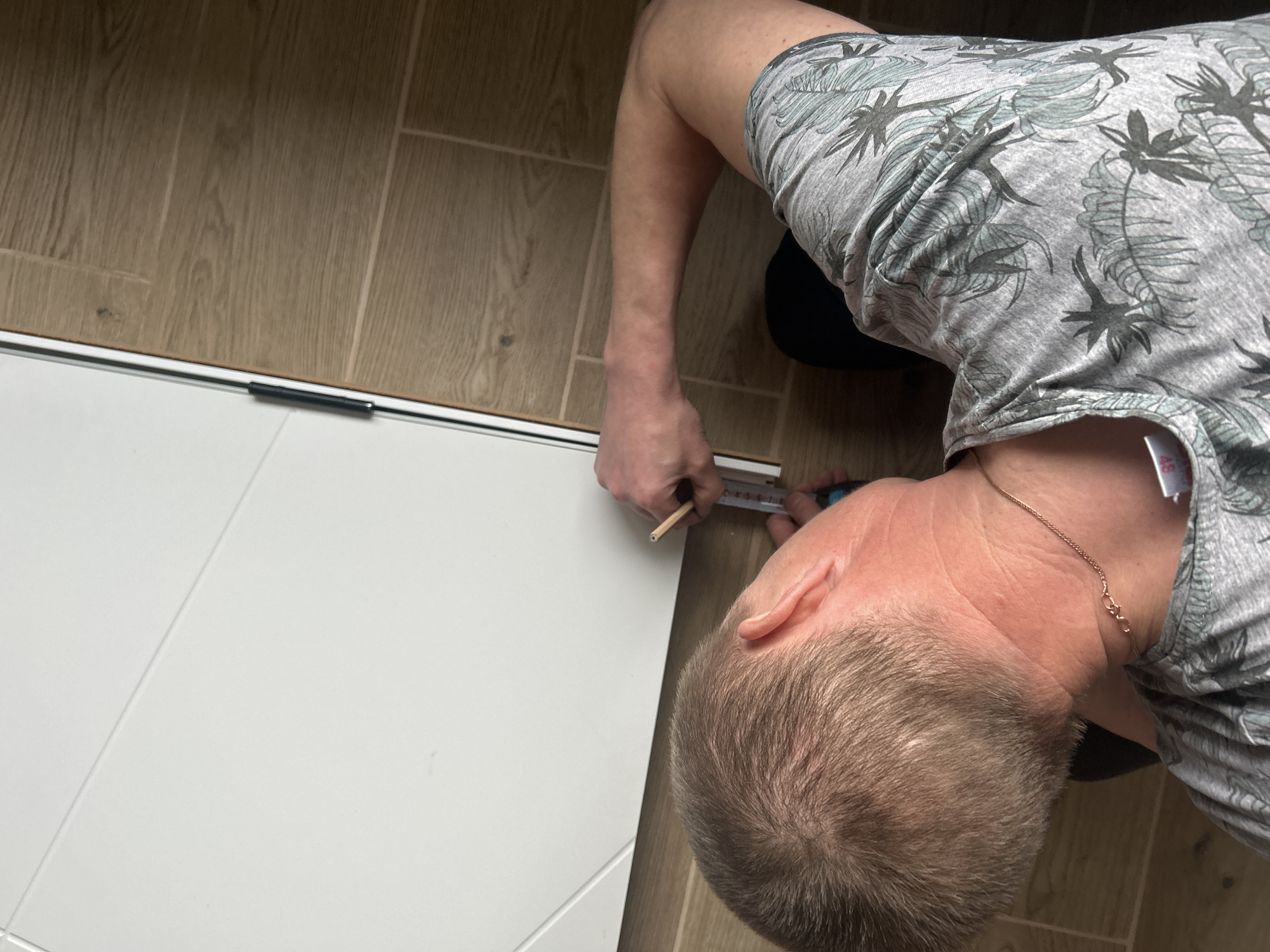 A top-down shot depicts a man measuring a white door with a measuring tape and a pencil. He's kneeling on the floor, which is covered in light brown wood-look planks. The man is wearing a gray t-shirt with a palm tree pattern and a gold chain around his neck. A small white tag is visible on his shirt. The location is Chaikovsky, Russia.