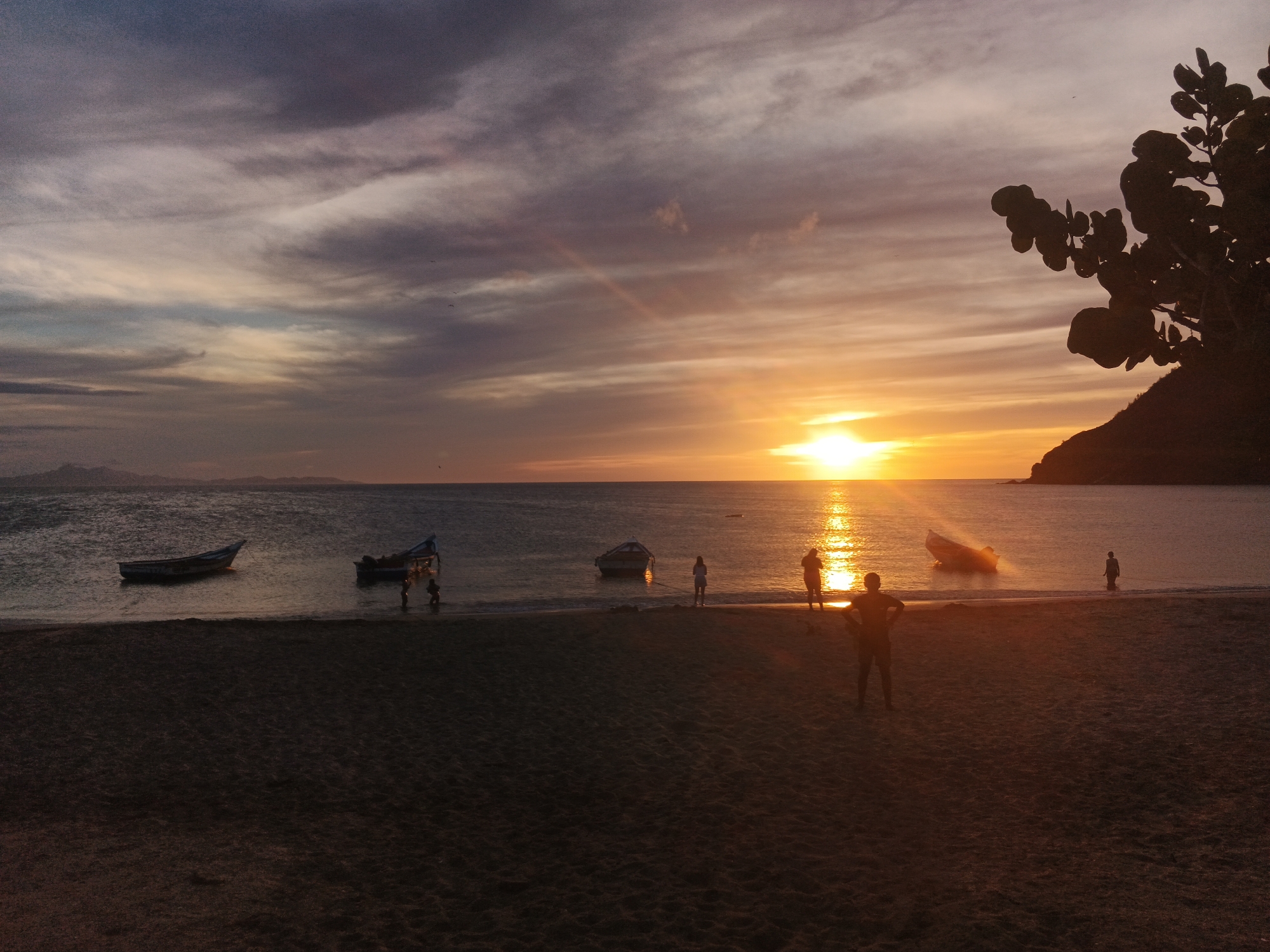 This is a wide shot of a beach at sunset. The sky is a mix of purple, orange, and yellow clouds, with the sun setting on the horizon. The ocean is calm and reflects the colors of the sky. Several boats are floating in the water near the shore. In the foreground, there is a sandy beach with several silhouettes of people. One person stands in the center of the frame, facing away from the camera, with their hands on their hips. Other figures are scattered across the beach and in the shallow water. To the right of the frame, a silhouette of a tree and a hill are visible against the sky. The overall mood of the image is peaceful and serene.