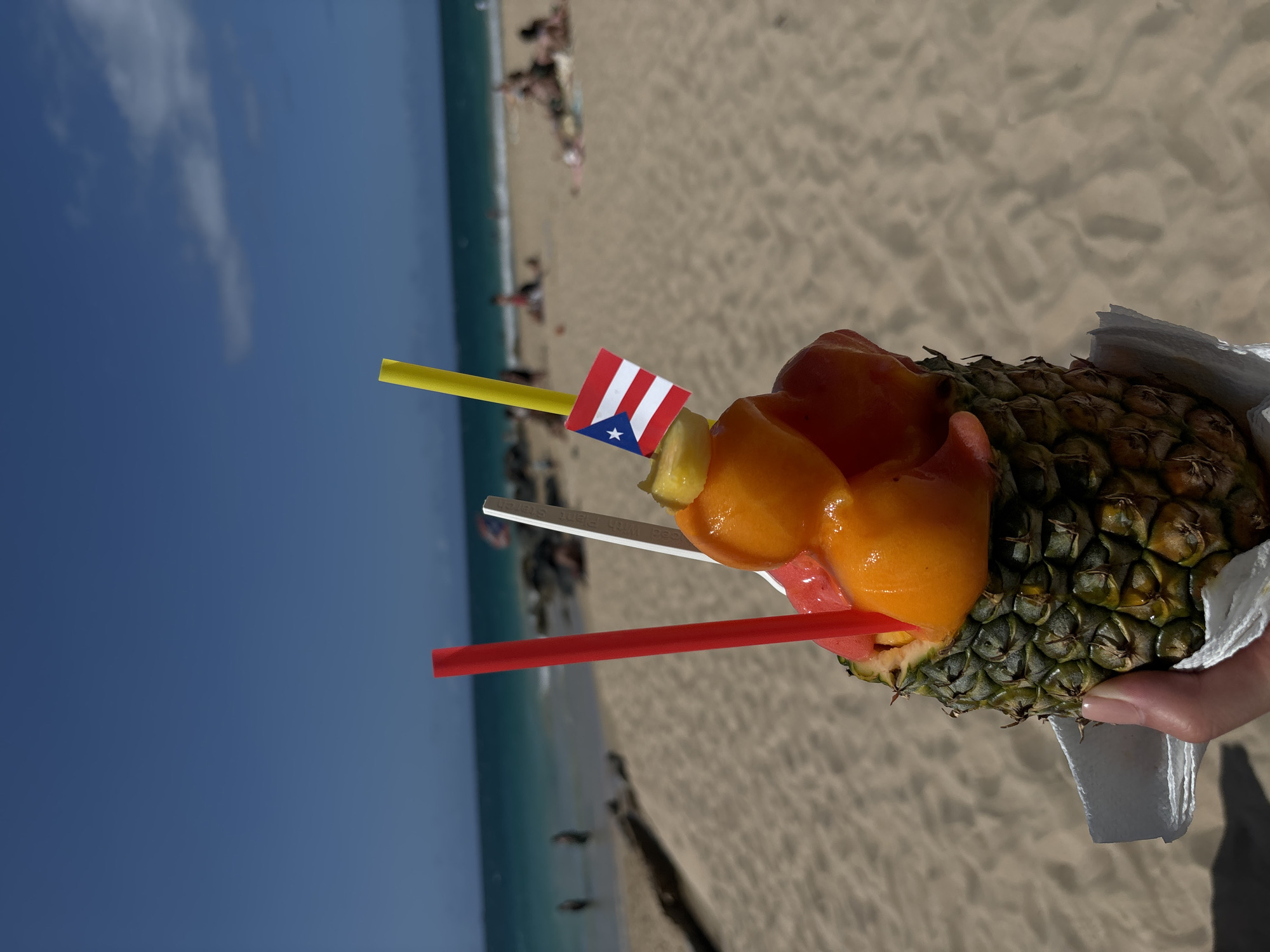 A hand holds a hollowed-out pineapple filled with colorful scoops of fruit, possibly sorbet or ice cream, and topped with a small Puerto Rican flag. The pineapple is presented against a blurred background of a sandy beach with turquoise ocean water and a clear blue sky. Several people are visible in the distance, some sunbathing or walking along the shore. The scene suggests a tropical vacation setting, likely in San Juan, Puerto Rico.