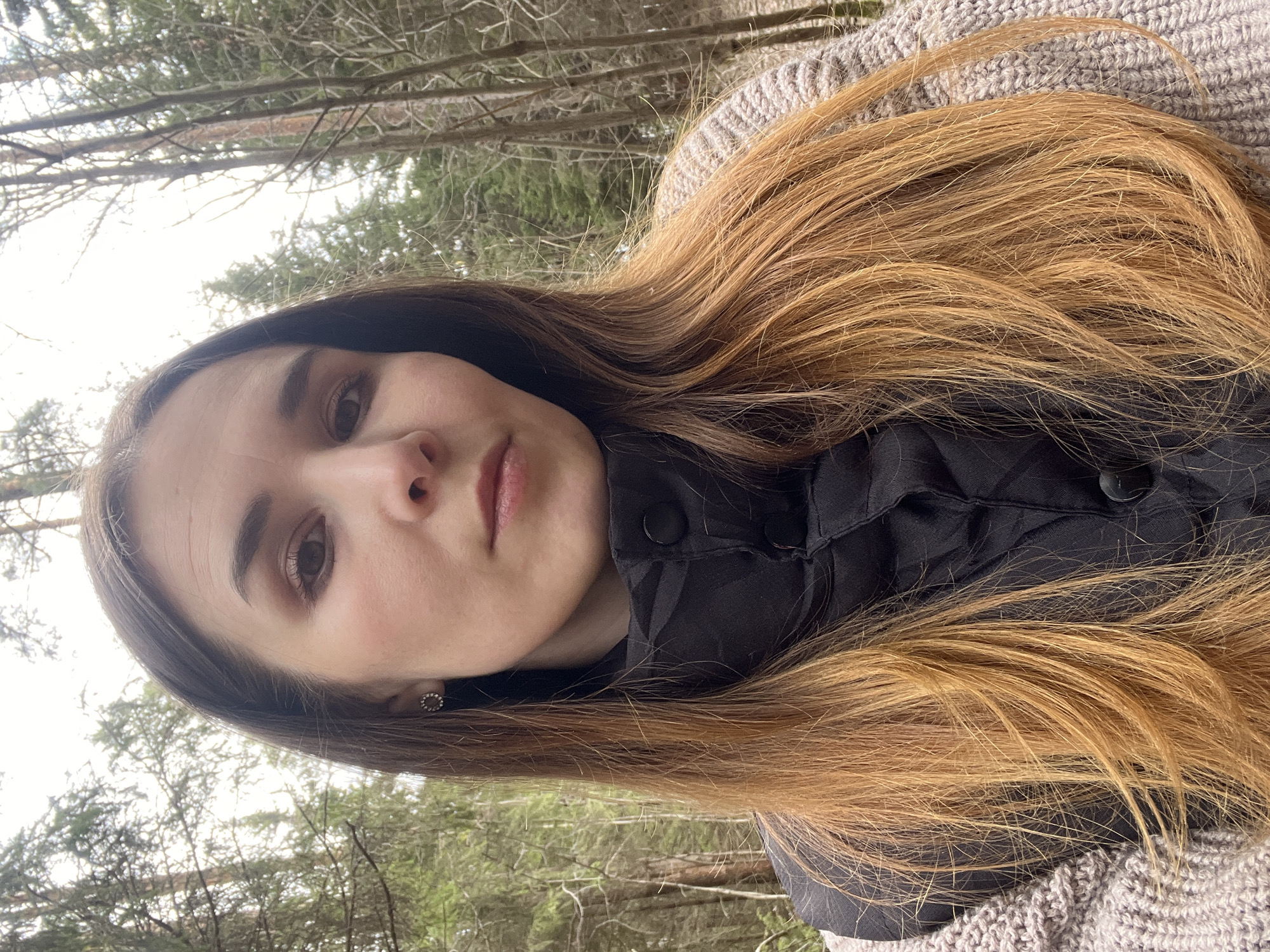 A portrait features a young individual, likely female, captured outdoors in what appears to be a forest or wooded area in Камское, Russia. The image is oriented with the subject's head tilted, appearing to look upwards towards the camera, with the background trees above her.
The subject has long, flowing hair that is a prominent golden-brown or light auburn color, cascading over her shoulders. She has a neutral expression, with visible brown eyes and dark eyebrows. She is wearing a dark, possibly black, button-up jacket or vest, which features a subtle, almost plaid-like pattern on the fabric. A light grey, chunky knit garment, possibly a sweater or scarf, is visible around her shoulders and neck, contrasting with the dark jacket. A small, circular, dark-colored earring is visible in her right ear.
The background consists of numerous bare tree branches against a bright, overcast sky, suggesting a cool climate, possibly late winter or early spring before foliage has fully appeared. The lighting is diffused and bright, indicative of a cloudy daytime. No specific activity is depicted beyond the portrait itself, likely a self-portrait or a close-up taken by another. There is no visible text in the image.