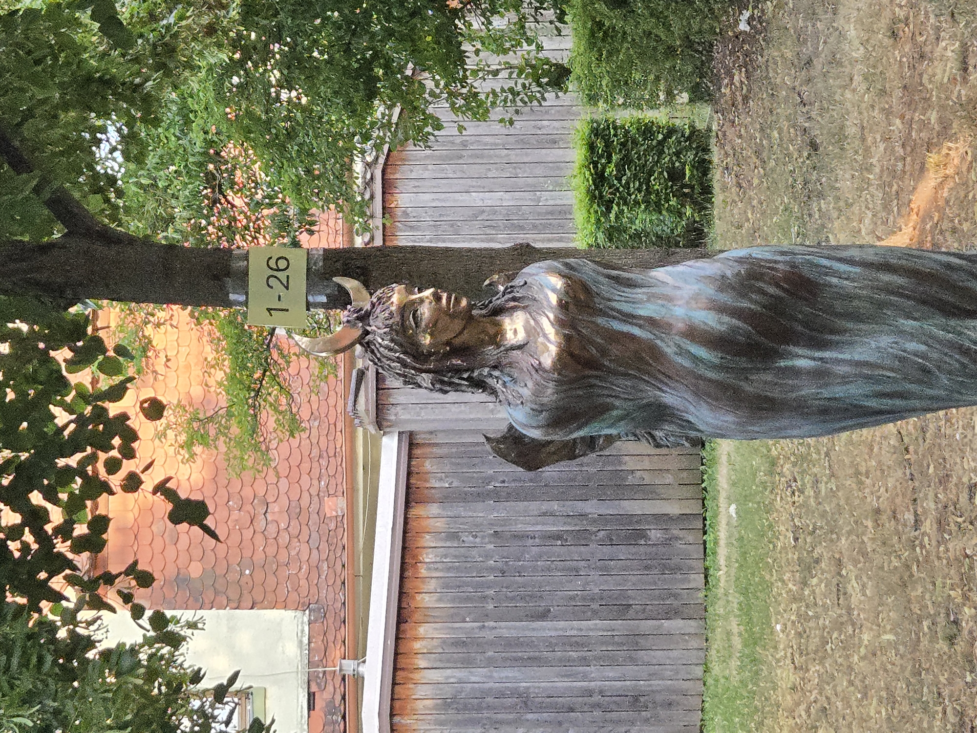 The image captures a statue of a woman with crescent horns on her head, resembling a pagan deity, standing in a garden setting. The statue is made of bronze and has a flowing garment. Behind the statue, a wooden fence runs horizontally, and a tree trunk is visible with a sign attached to it that reads "1-26". The background shows a building with red-tiled roofing and lush green foliage. The lighting suggests it is daytime, with sunlight illuminating the scene. This statue is located in the Hofgarten in Ansbach, Germany, near the Orangerie.
