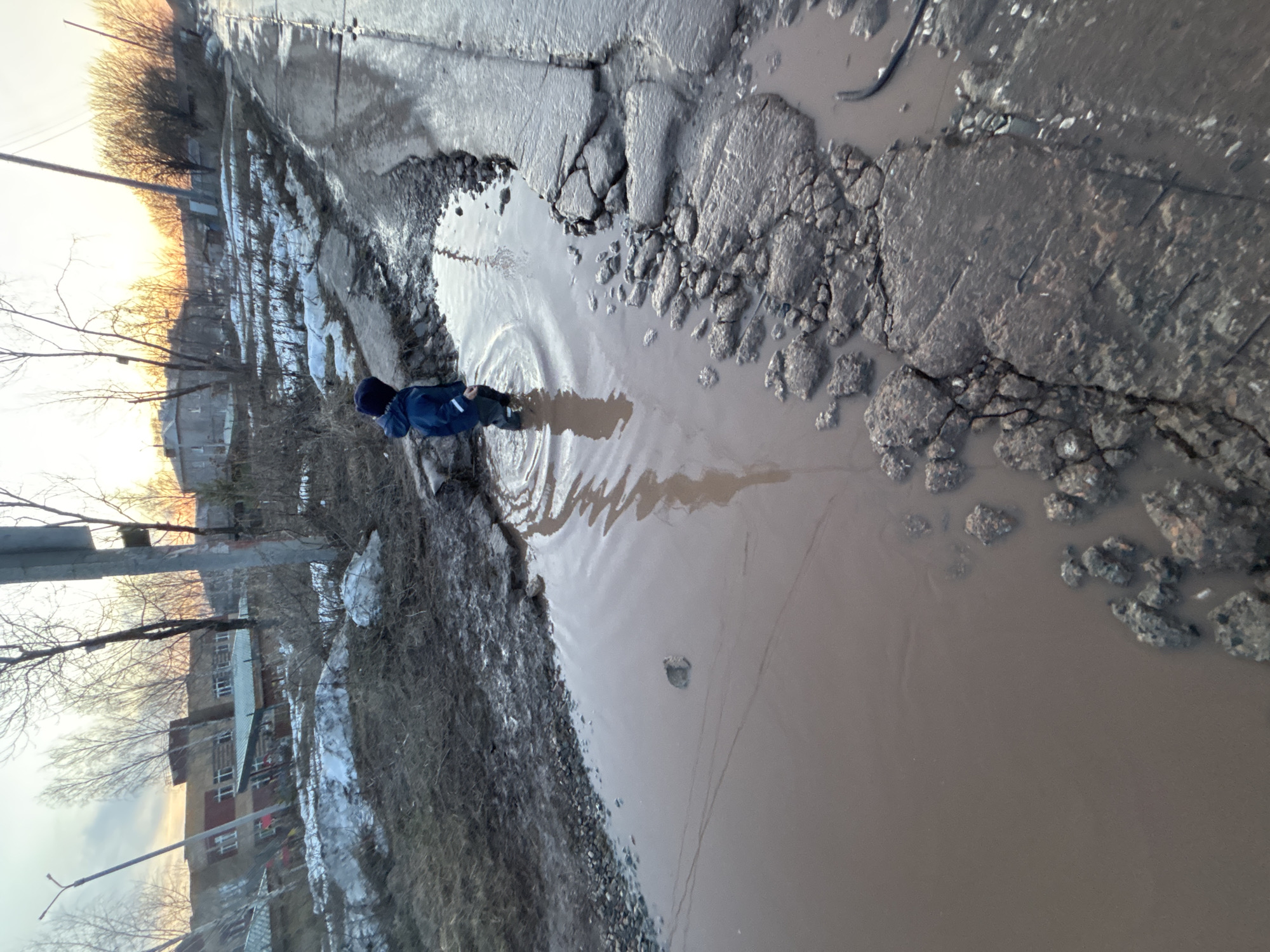 The image shows a waterlogged and damaged road in the city of Ферма, Russia. A young person wearing a blue jacket and dark pants is walking through a large puddle, causing ripples in the water. The road is severely cracked and broken, with chunks of asphalt missing. Melting snow and ice are visible along the roadside, indicating a transition from winter to spring. Buildings and bare trees are in the background, silhouetted against a bright, possibly setting, sun. A utility pole stands on the left side of the frame. The water in the puddle is murky and brown.