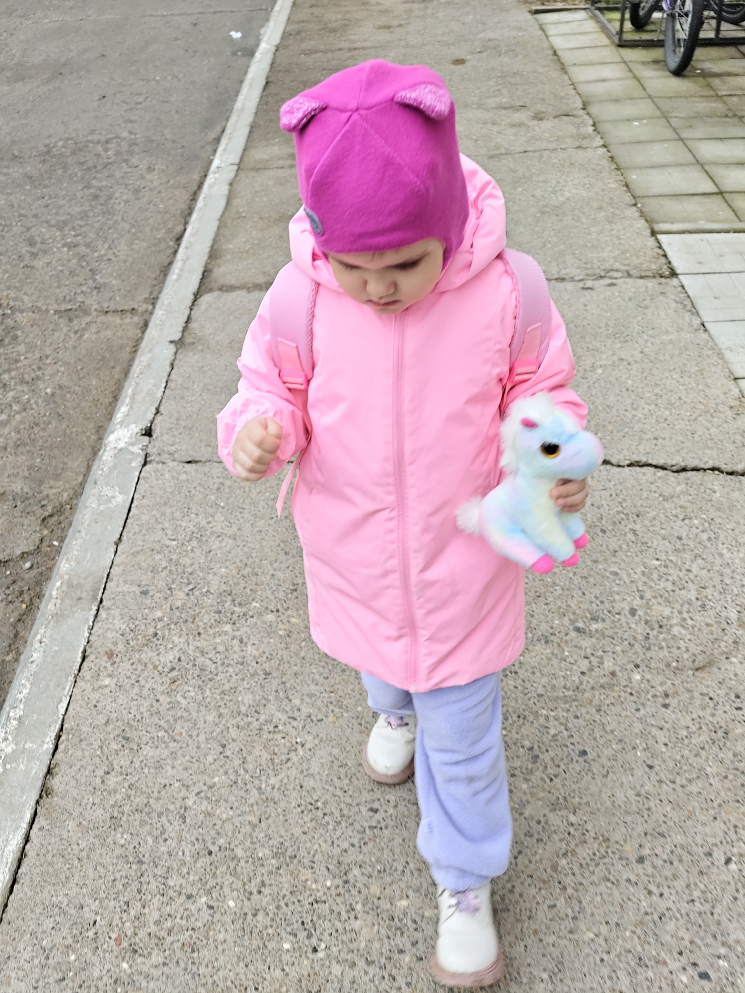 A young child wearing a bright pink winter coat and a matching pink hat with ears is walking on a concrete sidewalk. They are carrying a small, plush unicorn toy. The child is also wearing light purple pants and white shoes with pink soles. The sidewalk is cracked in places and borders a curb on one side and a paved area on the other. In the background, bicycles are parked on a similar concrete surface. The location is most likely Chaikovsky, Russia.
