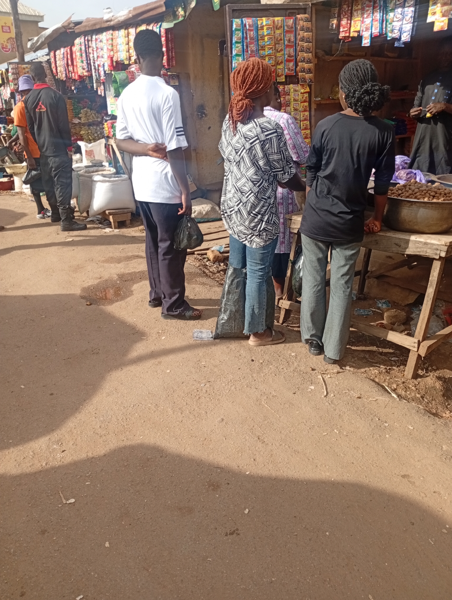 A medium shot captures a street market scene with at least six people, viewed from behind and slightly to the side. The market appears to be in an open outdoor setting with a dirt ground. In the background, a stall displays various packaged goods hanging in rows. To the left of the frame, three individuals are partially visible. One person wears an orange t-shirt and a purple headscarf. Next to them, a person in a black long-sleeved shirt with a red logo and black pants stands facing the stall. Another person, in a white t-shirt and dark pants, stands to the right of these two. In the center of the image, two individuals are more prominent. One person wears a patterned black and white top and blue jeans, with their hair styled in reddish braids. Beside them, another person is dressed in a black long-sleeved shirt and grey pants. To the far right, partially obscured, is a person in dark clothing standing behind a wooden stall. This stall has a metal bowl filled with nuts on top. The ground is dusty and uneven, with some shadows cast by an unseen object or structure. A puddle of water is visible on the ground in the foreground. The general atmosphere suggests a bustling, everyday marketplace.
