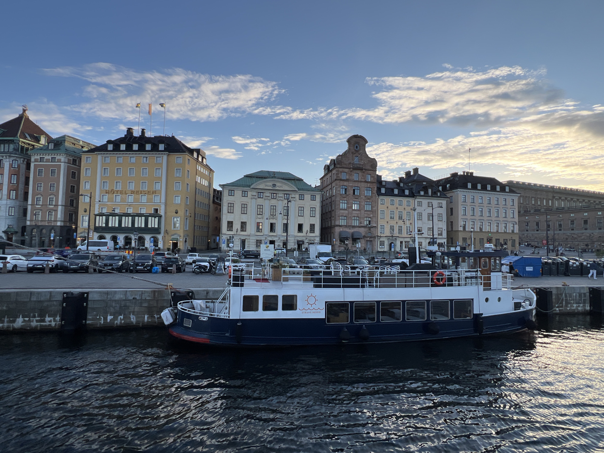 The image depicts a waterside scene in Stockholm, Sweden, likely in the central part of the city given the architecture. A white and dark blue passenger boat is docked at a quay. The boat has several windows along its side and a white railing. A life preserver is visible on the deck. Behind the quay, a row of multi-story buildings lines the waterfront. These buildings exhibit a variety of architectural styles, with some featuring classical elements and others a more modern design. The facade of one prominent building displays the sign "HOTEL REISEN." Flags are flying from poles on top of some buildings. In the background, cars and a van are parked along the street adjacent to the quay. The sky above is partly cloudy, with sunlight breaking through, suggesting it is either early morning or late afternoon. The water in the foreground is rippling, reflecting the light and the colors of the sky and buildings.
