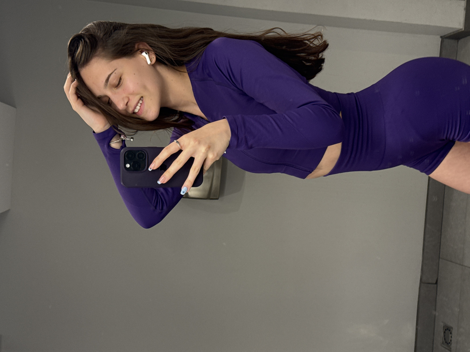 A young woman with long brown hair is taking a selfie in a bathroom. She is wearing a purple, form-fitting athletic outfit consisting of a long-sleeved top and shorts. She has wireless earbuds in her ears and is holding a black smartphone. The bathroom has gray walls and light gray tiles. The location is unidentifiable as Sofia, Bulgaria.