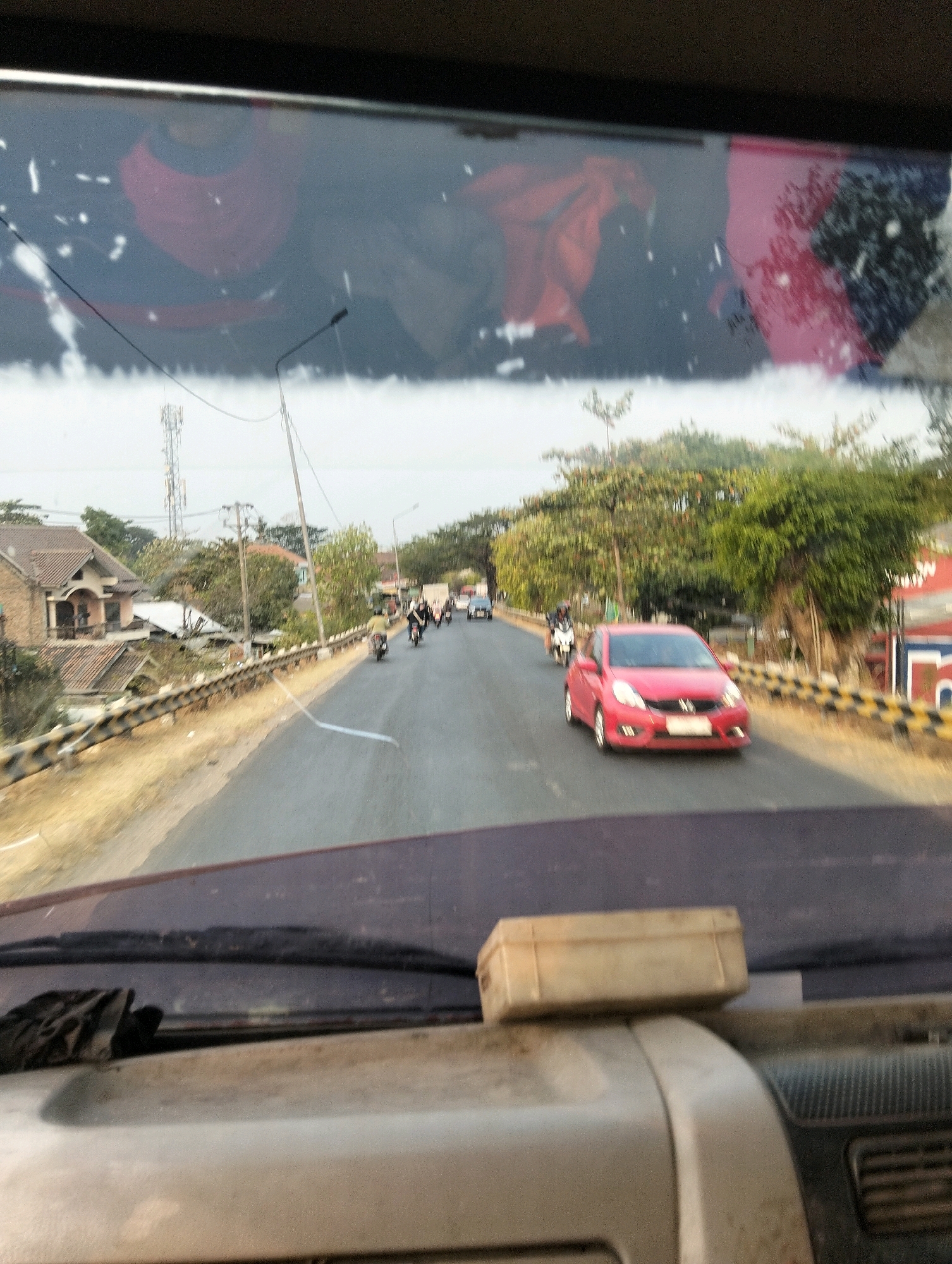 The image depicts a street scene in Semarang, Indonesia, viewed from inside a vehicle, likely a truck given the dashboard and windshield wiper visible at the bottom. The time of day appears to be daytime, with overcast skies contributing to diffused lighting. The road is paved and two lanes wide, with a metal guardrail separating the road from a dry, grassy embankment on the left.
Several vehicles are present. In the foreground on the right, a red hatchback is driving on the road. Further ahead, multiple motorcycles are visible, with some riders appearing to be male. A few other cars are also seen in the distance.
The environment includes residential buildings with tiled roofs on the left side of the road, interspersed with trees and vegetation. Utility poles and communication towers are also present, indicating an urban or semi-urban setting. The reflection on the windshield shows blurry figures, possibly passengers or items within the vehicle, with patches of red and pink colors. The windshield itself appears to have some dirt or smudges.
A small, light-colored rectangular object rests on the dashboard in the lower right portion of the frame, possibly a storage container or similar item. A dark, fabric-like object, possibly a cloth or glove, is visible on the left side of the dashboard.