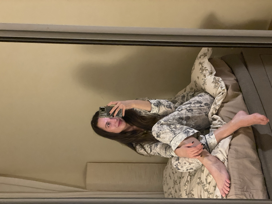 A woman in floral pajamas is reflected in a mirror. She sits on a bed with a beige comforter and a patterned pillow. She holds a phone up, presumably taking a selfie. The background features a tan wall, and a headboard is partially visible. The mirror's frame is silver, providing a defined border to the scene. The woman has long, brown hair. Her toes are pointed upward, and her legs are crossed.