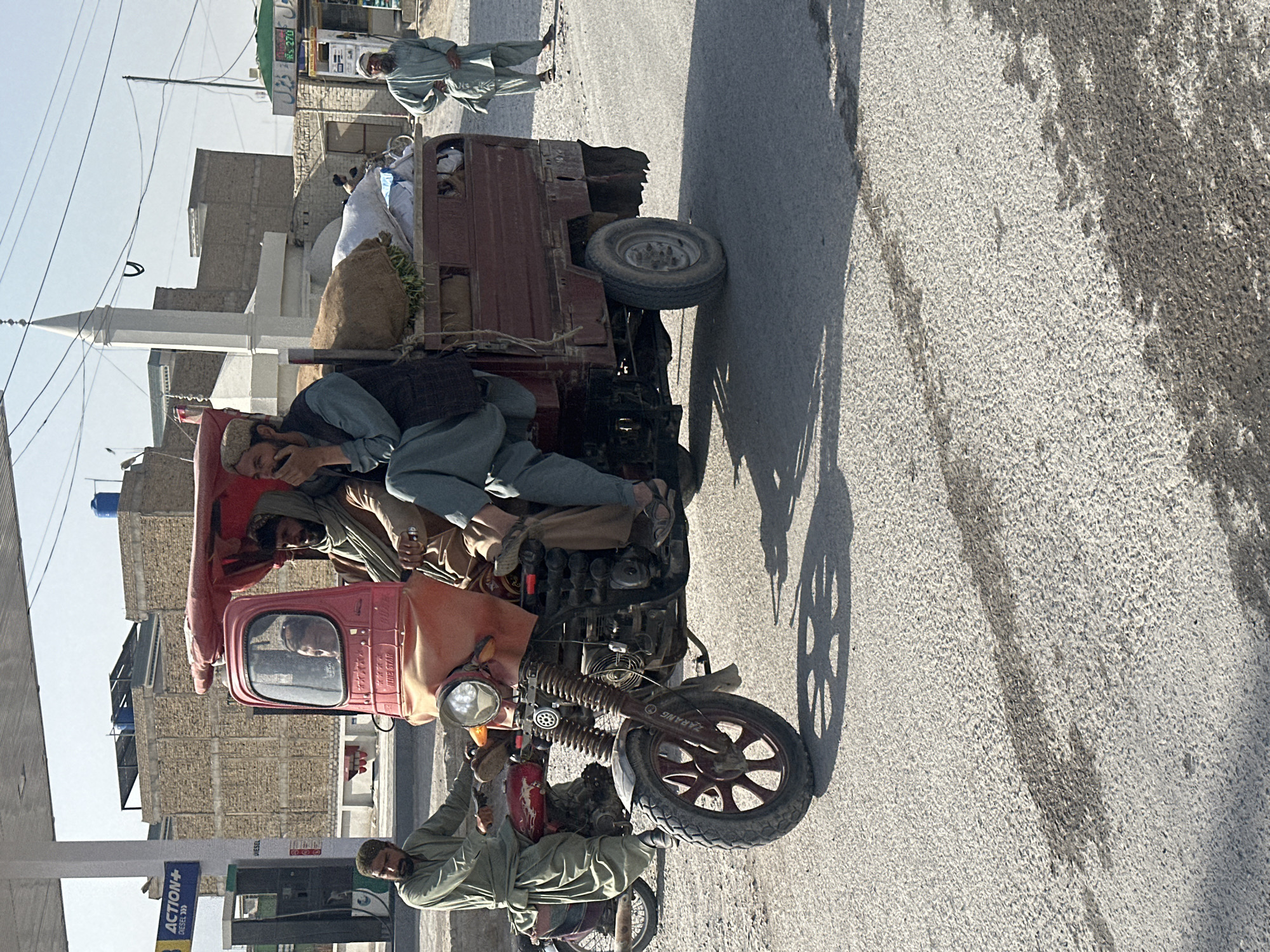 The image captures a roadside scene in Quetta City Tehsil, Pakistan, presenting a three-wheeled vehicle loaded with goods and carrying several individuals. A man in traditional Pakistani clothing sits atop the loaded vehicle, seemingly engaged in a phone conversation. Another man stands near the front of the vehicle.
In the background, buildings typical of urban Pakistan form the backdrop, and a mosque minaret is visible. The weather appears sunny and clear. The road is paved and shows signs of wear and tear. A petrol station sign is seen in the left corner of the image.