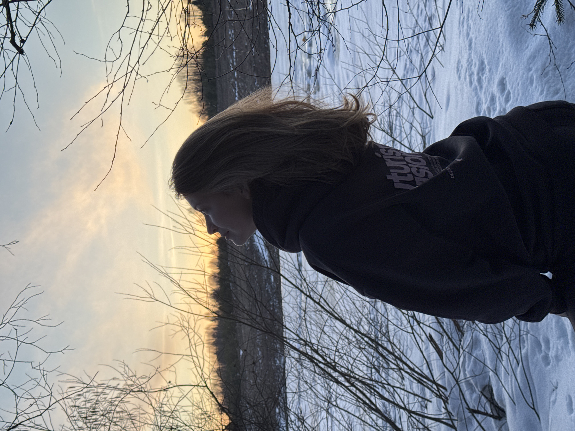 A photograph shows a woman from the side, facing left, in an outdoor setting in Вассята, Russia. The backdrop consists of a winter landscape, with snow-covered ground and bare branches suggesting the season. The time of day appears to be around sunset. The woman is wearing a dark-colored hoodie. The surrounding vegetation consists of dormant trees and shrubs.