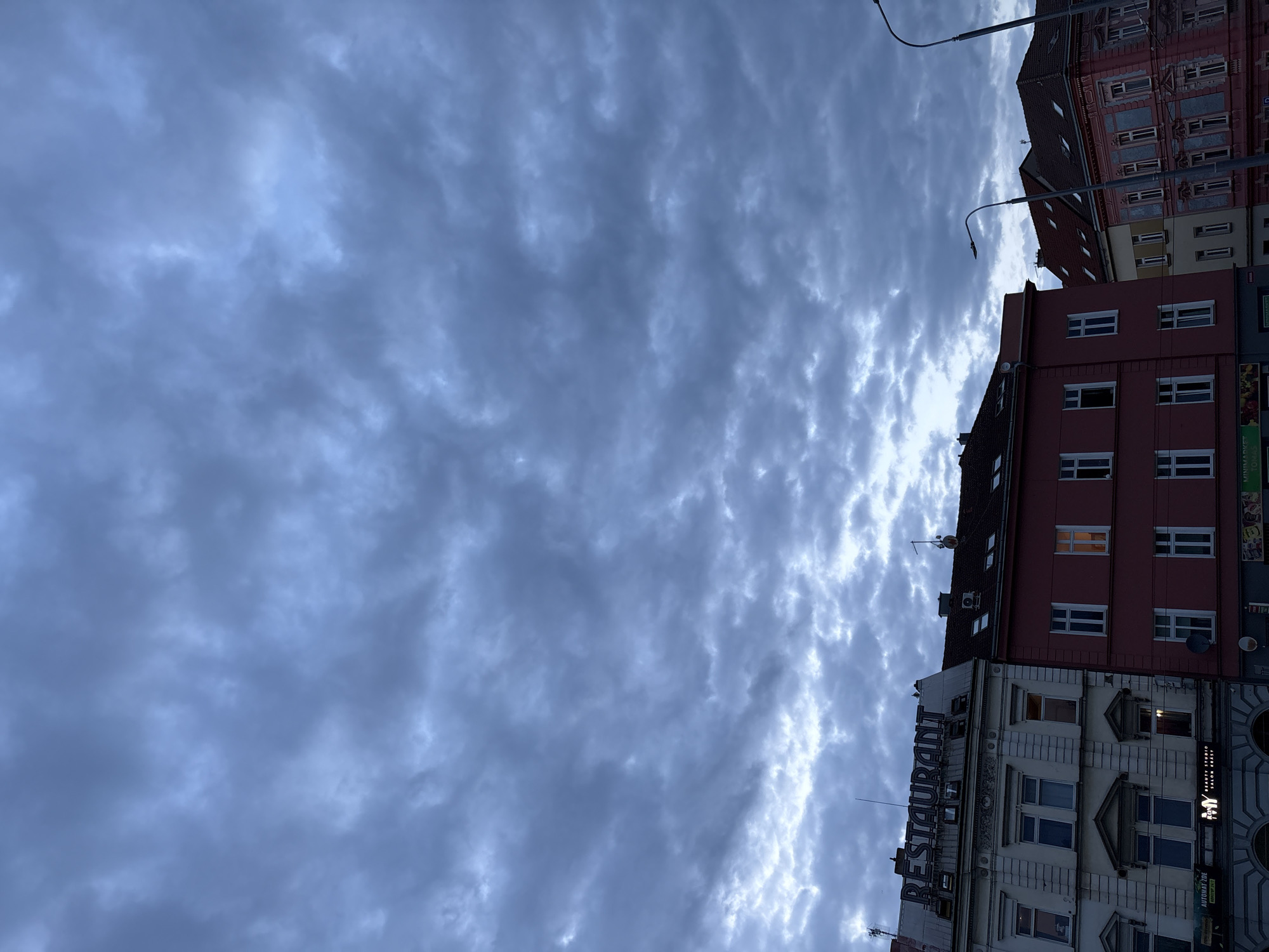 The image shows a street view in the city of Pilsen, Czechia, under a cloudy sky. The foreground is dominated by the facades of several buildings, with a prominent "RESTAURANT" sign on the leftmost building. The buildings are multi-story structures with various architectural styles and colors, including white, pink, and light yellow. Windows and some illuminated interiors are visible. In the background, two streetlights are present. The sky is filled with a dense layer of dark, textured clouds. No people are clearly visible in the image.