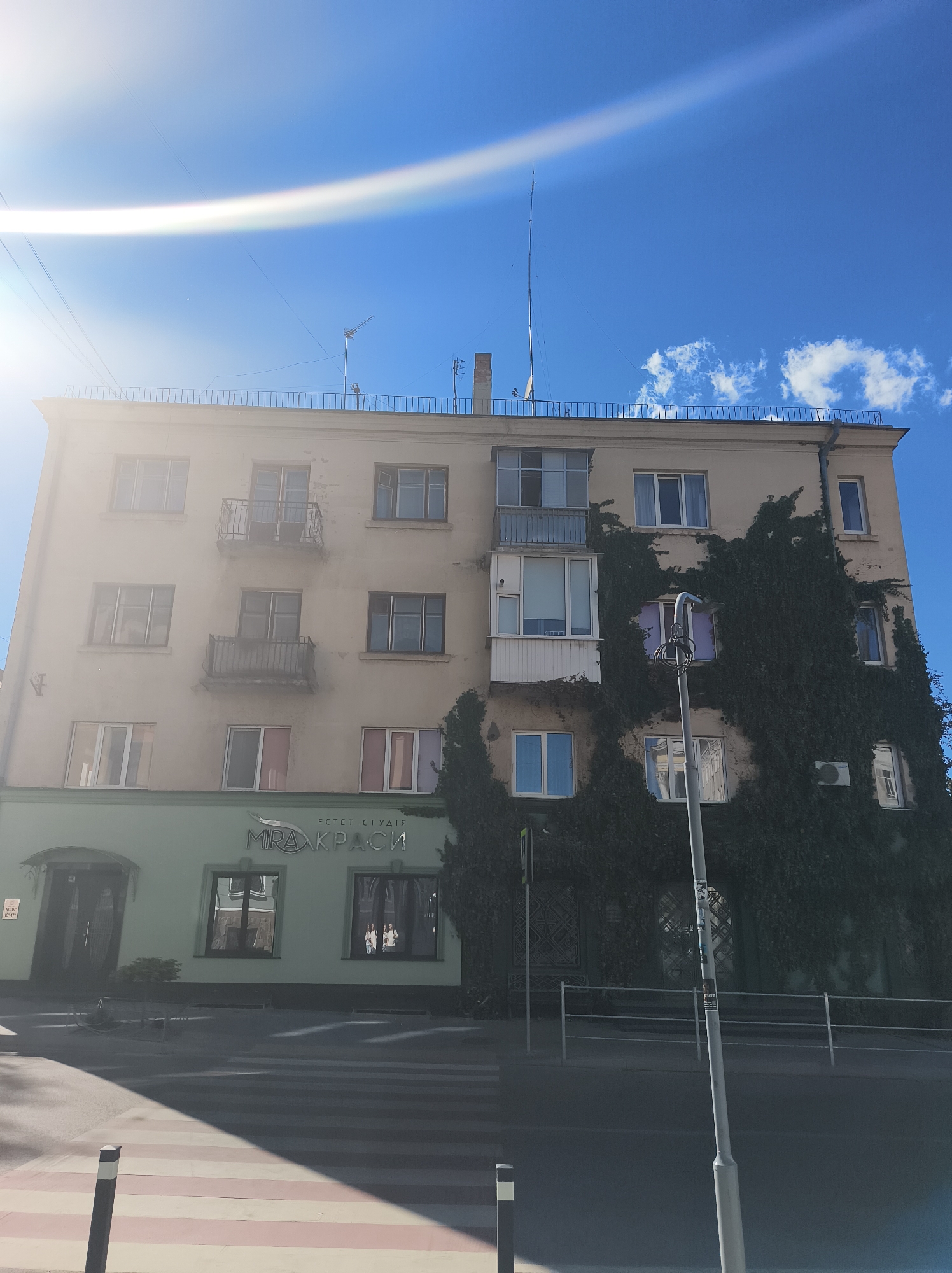 The image shows a four-story building in Ternopil, Ukraine. The building has a light-colored facade and a flat roof with a railing. The left side of the building has balconies, while the right side is covered in ivy. The lower part of the building has a light green storefront with windows and a sign that reads "Естет Студія МІРА КРАСИ" (Estet Studio MIRA KRASI). A pedestrian crosswalk and street lighting are in front of the building. The sky is blue with some clouds and lens flare from the sun.