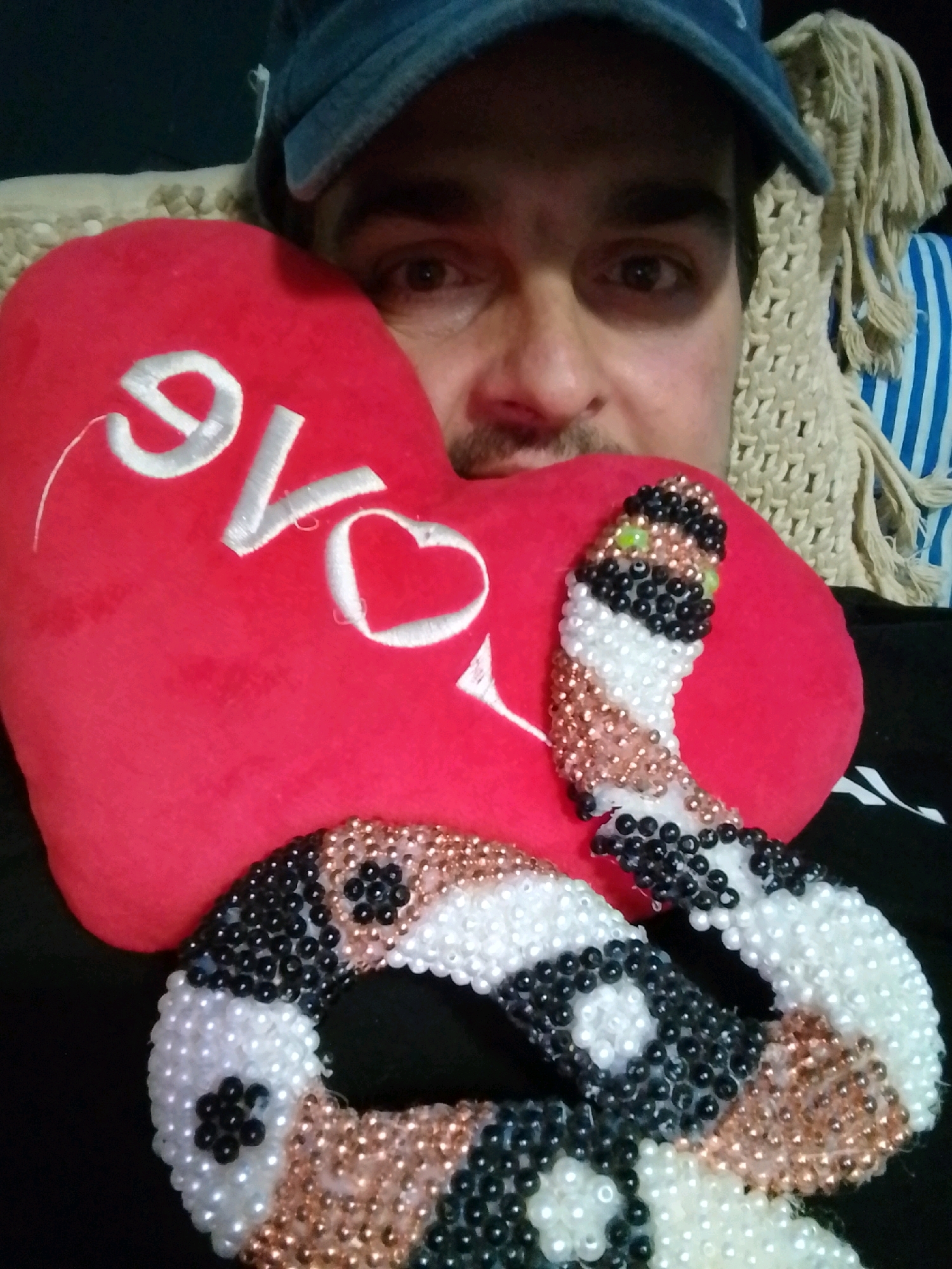 The image shows a close-up of a man wearing a blue baseball cap, partially obscuring his face. He is holding a red heart-shaped pillow with "LOVE" embroidered in white thread. Two beaded snake-like figures are placed in front of the pillow, one with green eyes. The background consists of a textured beige throw and a blue and white striped fabric. The man is lying down, and the image is taken from a high angle. The scene appears to be indoors, possibly a living room. There is no information to suggest the image is located in Wedel, Germany.