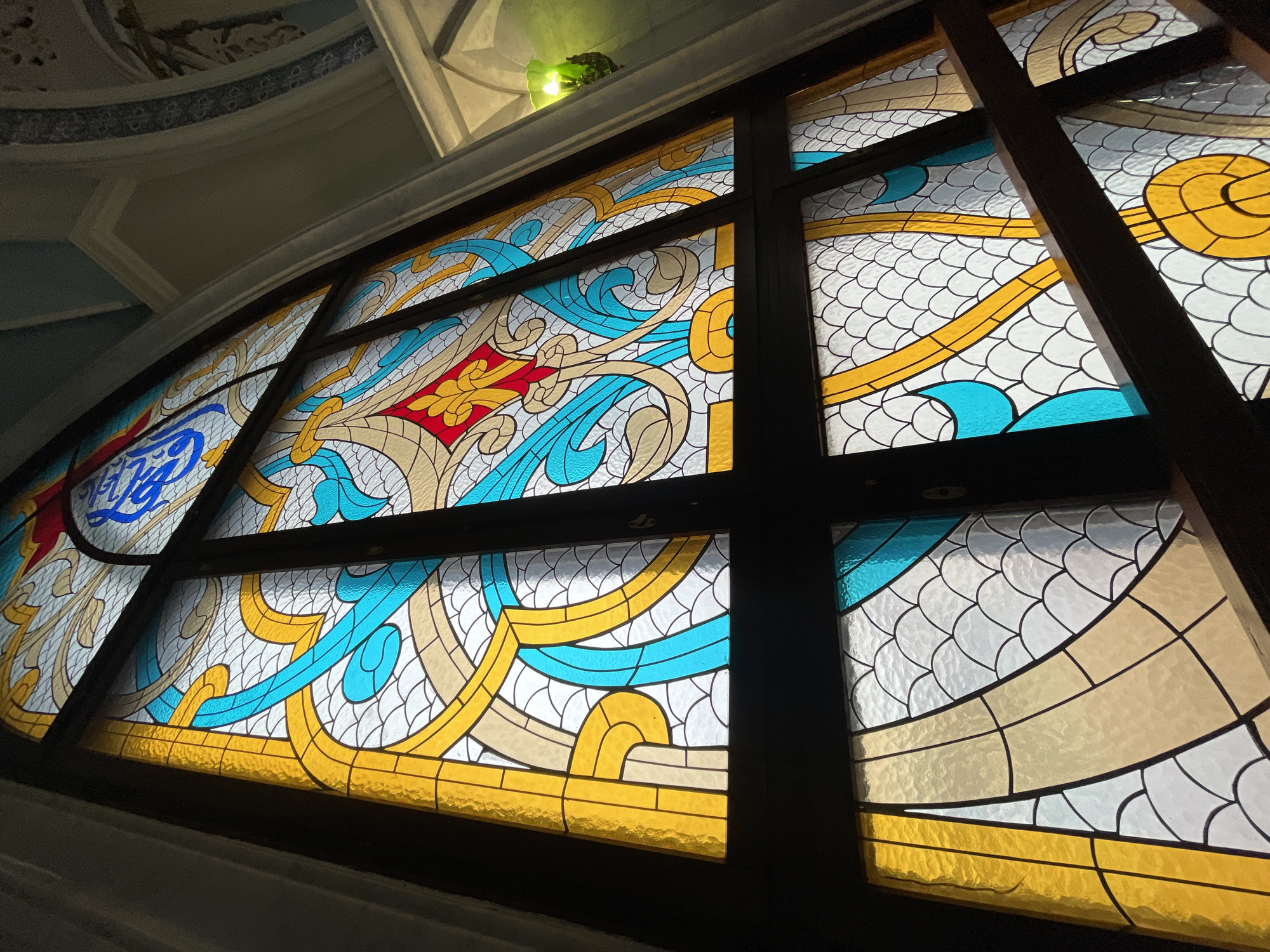 A low-angle, eye-level shot depicts a large, ornate stained glass window, its wooden frame dark brown and dividing the artwork into multiple panes. The window features a repeating pattern of teal blue and gold swirling designs against a background of white scales. The lower portion of the glass is yellow. On the left, a red and yellow floral design, and a blue emblem are visible. The window is presumably in Kazan, Russia. The architectural details of the building's ceiling are visible at the top of the frame.
