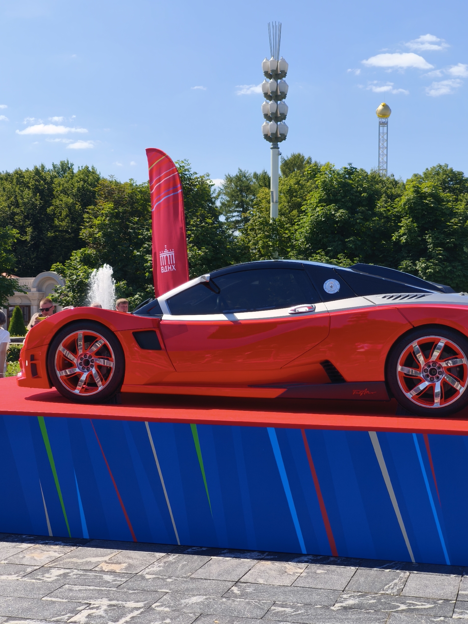 An eye-level, medium shot captures a vibrant red sports car displayed on a red platform against a backdrop of lush green trees and a clear blue sky in Moscow, Russia, likely at VDNKh, suggested by the iconic spire and the flag with Cyrillic text. The car, with its sleek design, black roof, and silver accents, is positioned slightly to the right of the frame. Its polished chrome wheels gleam under the sunlight, featuring intricate spoke designs.
In the background, a towering, multi-tiered light fixture, characteristic of VDNKh's architecture, stands prominently. Further in the distance, another tall, metallic structure is topped with a golden dome. A large red flag, partially obscured by the car, bears a graphic emblem and Cyrillic lettering, reinforcing the VDNKh location.
Several individuals are visible, appearing to admire the car. Two men are in the foreground, looking towards the vehicle, while others are partially visible to the left. A fountain with water jets is also present behind the car, adding a touch of dynamism to the scene. The foreground consists of a paved area with square, grey stone tiles, slightly wet or weathered, indicating an outdoor public space. The lower portion of the frame features a blue display panel decorated with abstract, colorful diagonal stripes in red, white, green, and blue. The overall impression is of a sunny day at an outdoor exhibition or event.
