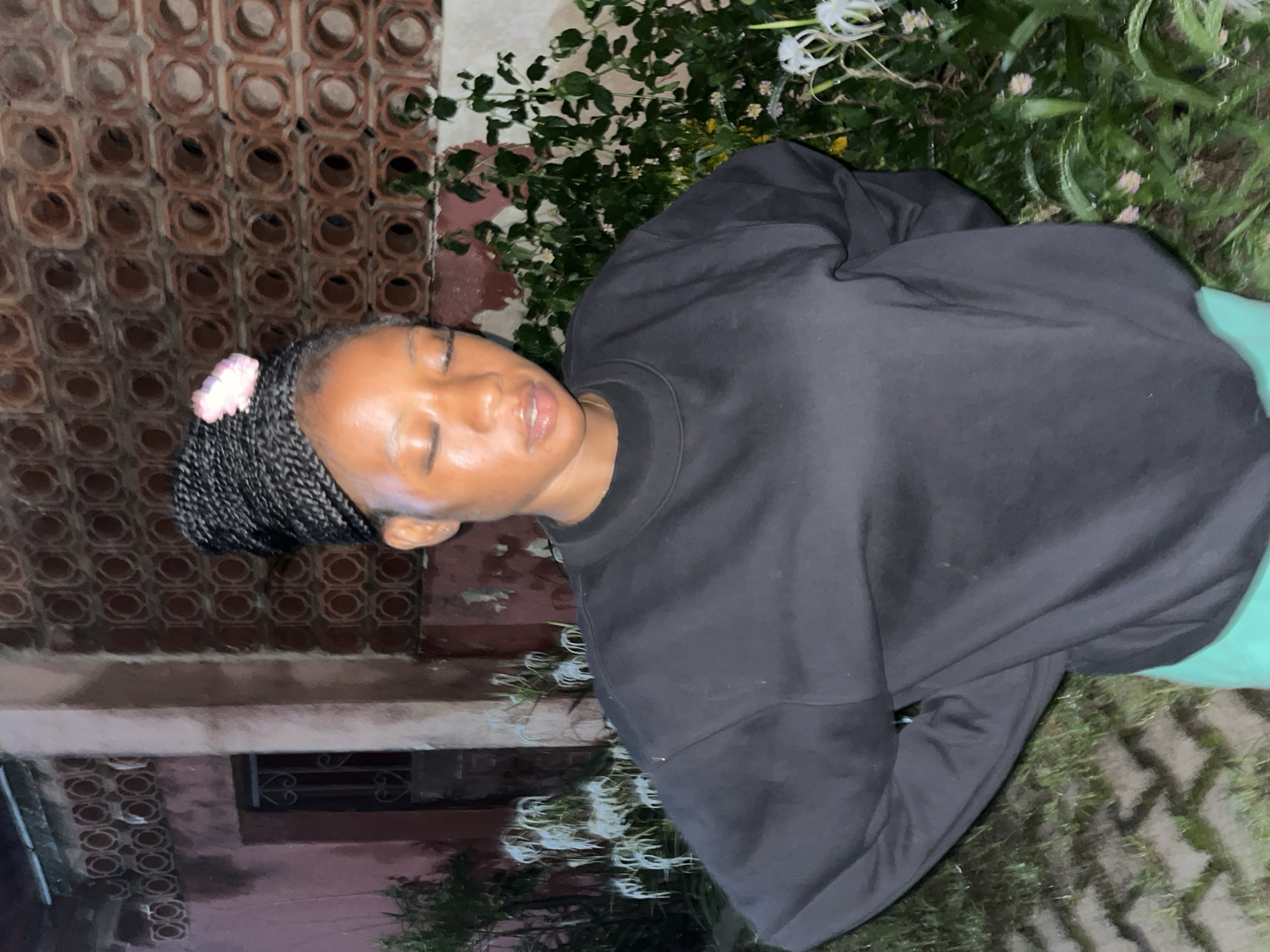 A young woman with braided hair, adorned with a pink flower, stands outside with her eyes closed. She is wearing a black sweatshirt and bright teal shorts. Behind her, a textured wall with circular cutouts and a closed metal gate is visible. Lush green foliage with white flowers is present to the right and left of the woman. The ground appears to be paved with interlocking bricks. This scene is located in Jos, Nigeria.