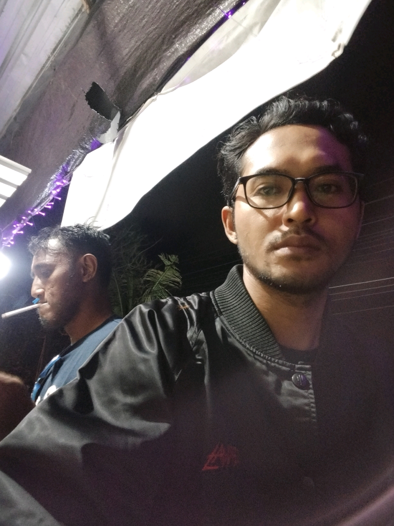 Two individuals are visible in this nighttime outdoor setting, likely in Denpasar, Indonesia, under a dark, tarp-like canopy that appears to have purple string lights hanging from it. In the foreground, a man with glasses and a black bomber jacket is facing the camera, his expression neutral. Behind him and to the left, another man is seen in profile, wearing a blue shirt and holding a lit cigarette to his mouth. The environment suggests a casual gathering or event.
