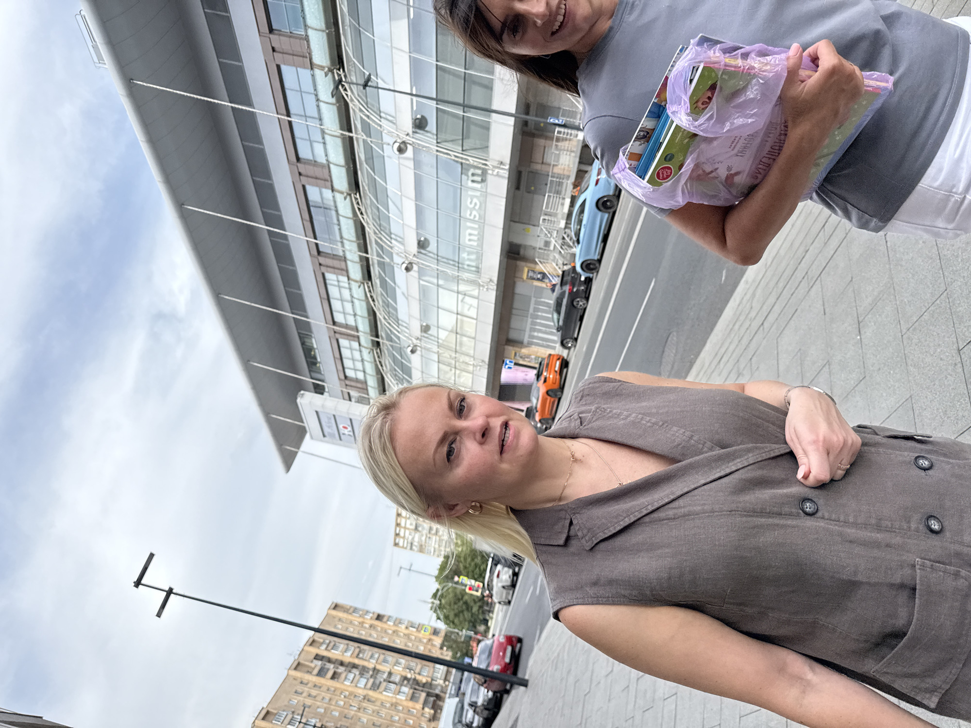 Two women are walking on a sidewalk in front of a modern building with a glass facade and a prominent sign that reads "mint missim". The woman on the left has blonde hair and is wearing a gray vest. She is looking towards the camera and speaking. The woman on the right has brown hair and is smiling. She is carrying a purple bag filled with books and magazines. In the background, cars are driving on the street, and several residential buildings can be seen. The sky is overcast. The scene appears to be a street in Moscow, Russia.