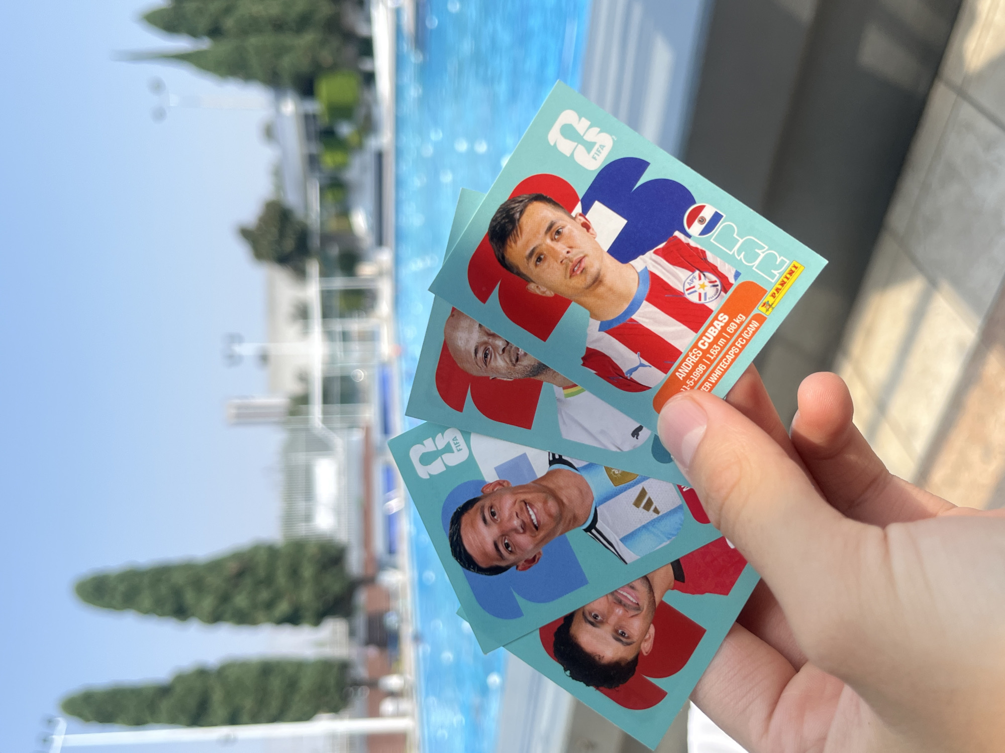 A hand holds three Panini FIFA World Cup 2022 sticker cards with pictures of soccer players. The top card depicts Andrés Cubas from Paraguay. Another card features a player from Argentina. The cards are held in front of an outdoor setting that includes a blue swimming pool and greenery, suggesting the cards are displayed near a recreational area.