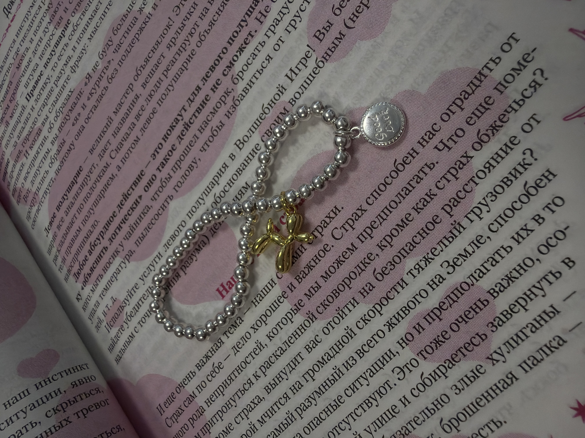 The image shows a close-up of an open book with a bracelet resting on its pages. The bracelet is made of silver beads and has a pendant with the inscription "Good Luck". A small, golden, balloon dog charm is also attached to the bracelet. The book's pages are covered in text and have a pink, heart-shaped pattern overlaying some of the text. The focus is on the bracelet and the book. There are no people or any identifiable locations within the city of Homyel, Belarus.