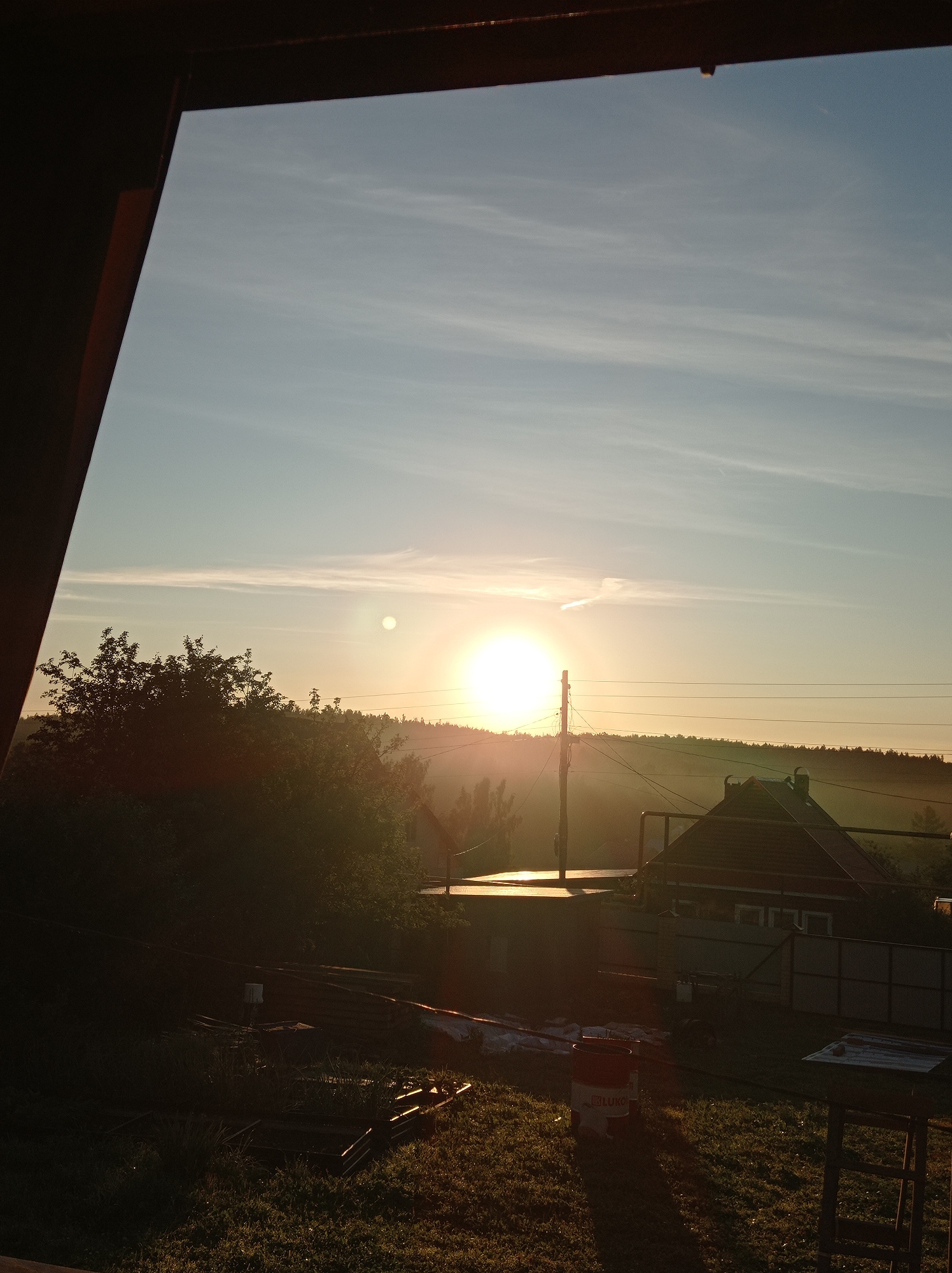 This is an outdoor, eye-level, medium shot photograph taken during sunrise. The scene depicts a rural or semi-rural environment, possibly on the outskirts of Degtyarsk, Russia, characterized by a clear sky with thin, wispy clouds. The sun is low on the horizon, casting a warm, golden light across the landscape.
In the foreground, a dark, angled structure, possibly a porch roof or overhang, partially frames the view. Below this, the ground is covered in grass and appears to be a yard or garden area. There are several objects scattered on the ground, including what appear to be metal frames for garden beds, some rolled-up material that could be tarpaulin or plastic sheeting, and a red and white bucket with writing on it, possibly a paint or chemical container. A wooden table or workbench is visible on the right side of the frame.
In the middle ground, a low-rise building with a dark, pitched roof and visible windows is situated. Adjacent to it is another flat-roofed structure. Trees and foliage are visible behind these buildings, forming a dark, silhouetted line against the brighter sky. A utility pole with multiple wires stretches across the scene horizontally.
The background consists of a gently rolling landscape covered in trees, suggesting a wooded or forested area that extends to the horizon. The overall impression is one of a peaceful, early morning scene in a quiet location. There are no people visible in the image.