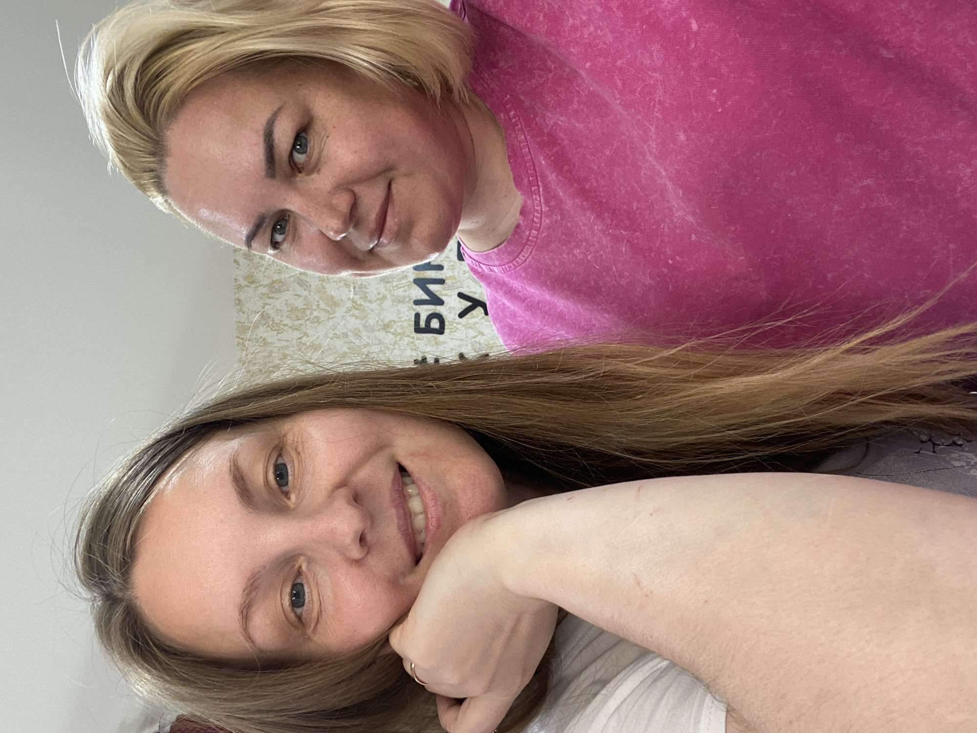Two women are in close proximity, appearing to be taking a selfie. The woman at the top has short blonde hair and is wearing a pink t-shirt. The woman at the bottom has long brown hair and is wearing a white t-shirt. The background is out of focus, but there is a wall with writing on it. The writing is in Cyrillic. There are no indications in the image to suggest this photo was taken in Noviy, Russia.