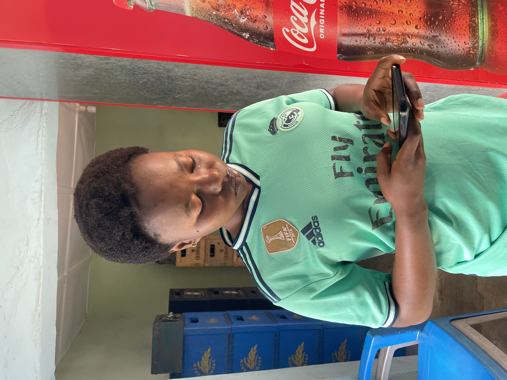 A young Black woman with short, dark, curly hair stands in what appears to be a small shop or convenience store in Jos, Nigeria. She is wearing a light green jersey with dark blue and white trim, featuring patches for "FIFA World Champions 2018" and "Adidas" on the left chest, and a Real Madrid crest on the right. The text "FLY PIRATES" is visible on the front of the jersey. She is holding a black smartphone in her hands, looking down at it, suggesting she is engaged with her device.
Behind her, stacked crates of beverages are visible. To her left, there are blue crates, some with the text "BREW" and a yellow motif. To her right, a large red Coca-Cola refrigerator is prominently featured, displaying condensation on the glass door which shows a large bottle of Coca-Cola. The background wall is a light green color. A blue plastic chair is partially visible in the bottom left corner of the frame. The lighting appears to be natural, coming from an unseen source, creating soft shadows. The overall impression is one of a casual, everyday setting within a commercial establishment.