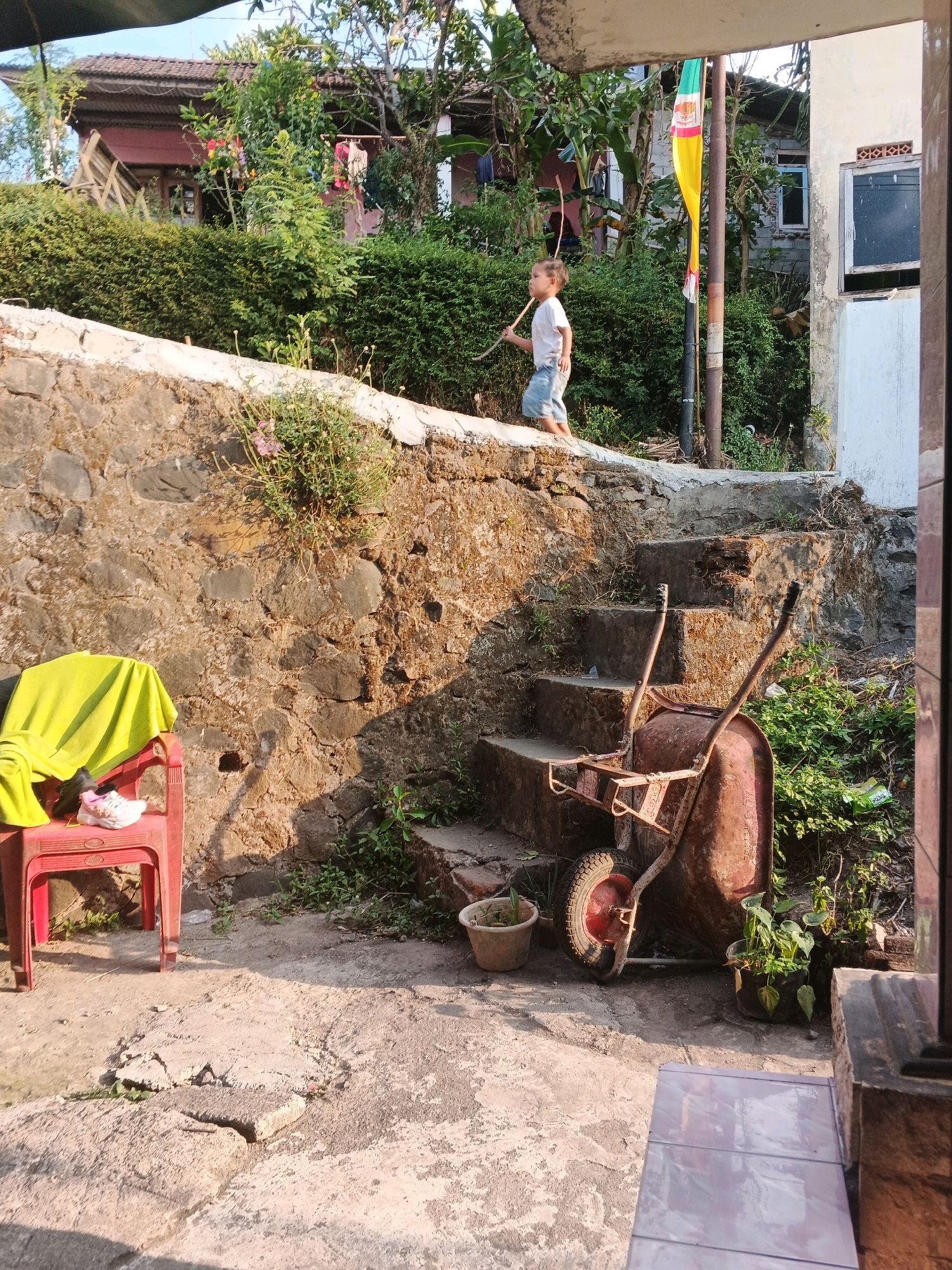 In Keji, Indonesia, a young boy, possibly of Indonesian descent, wearing a white t-shirt and blue shorts, walks along a stone wall, holding a stick. He is on the upper level of an outdoor area with a rustic ambiance. To his right, a weathered, rusty wheelbarrow stands on a set of concrete stairs. The stairs are surrounded by overgrown vegetation and a stone wall. In the foreground, a red plastic chair is draped with a bright yellow cloth and a pair of white sneakers. The scene appears to be a residential area with a building visible in the background, partially obscured by trees and foliage. A flag with green, yellow, and white colors is attached to a pole near the boy. The lighting suggests it is daytime, with sunlight casting shadows.