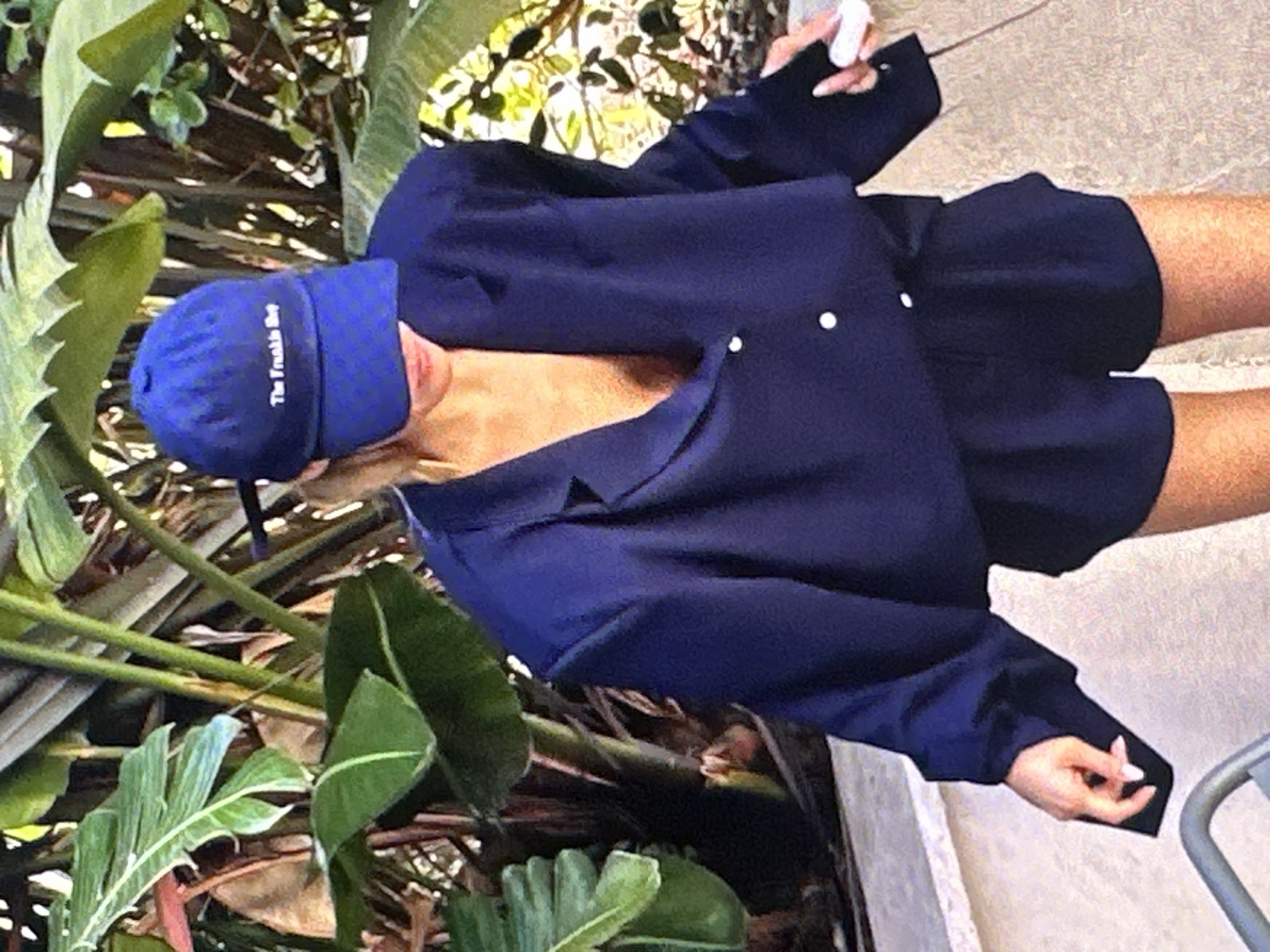The image depicts a person wearing a blue baseball cap with the text "The Frankie Boy" and a dark blue blazer-and-shorts outfit. The person is standing outdoors near lush green foliage, including banana leaves. A small portion of a gray object, possibly a metal chair or fence, is visible in the lower right corner. The location cannot be determined to be Yerevan, Armenia based on the provided image.