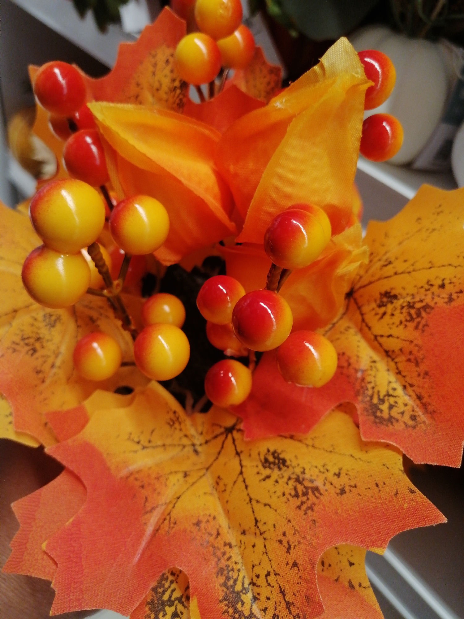 A close-up, eye-level shot features a cluster of artificial fall-themed decorative elements, primarily consisting of red and yellow berries and artificial maple leaves. The berries are small, round, and have a glossy finish, with some showing a gradient from red to yellow. They are clustered on thin, dark stems. The leaves are a vibrant orange and yellow, with some exhibiting dark speckles resembling veins or blemishes. The texture of the leaves appears to be fabric-like. The background is out of focus, but suggests an indoor setting with hints of white and gray objects, possibly shelves or furniture. There is no discernible indication of the specific location being Kremenchuk, Ukraine.