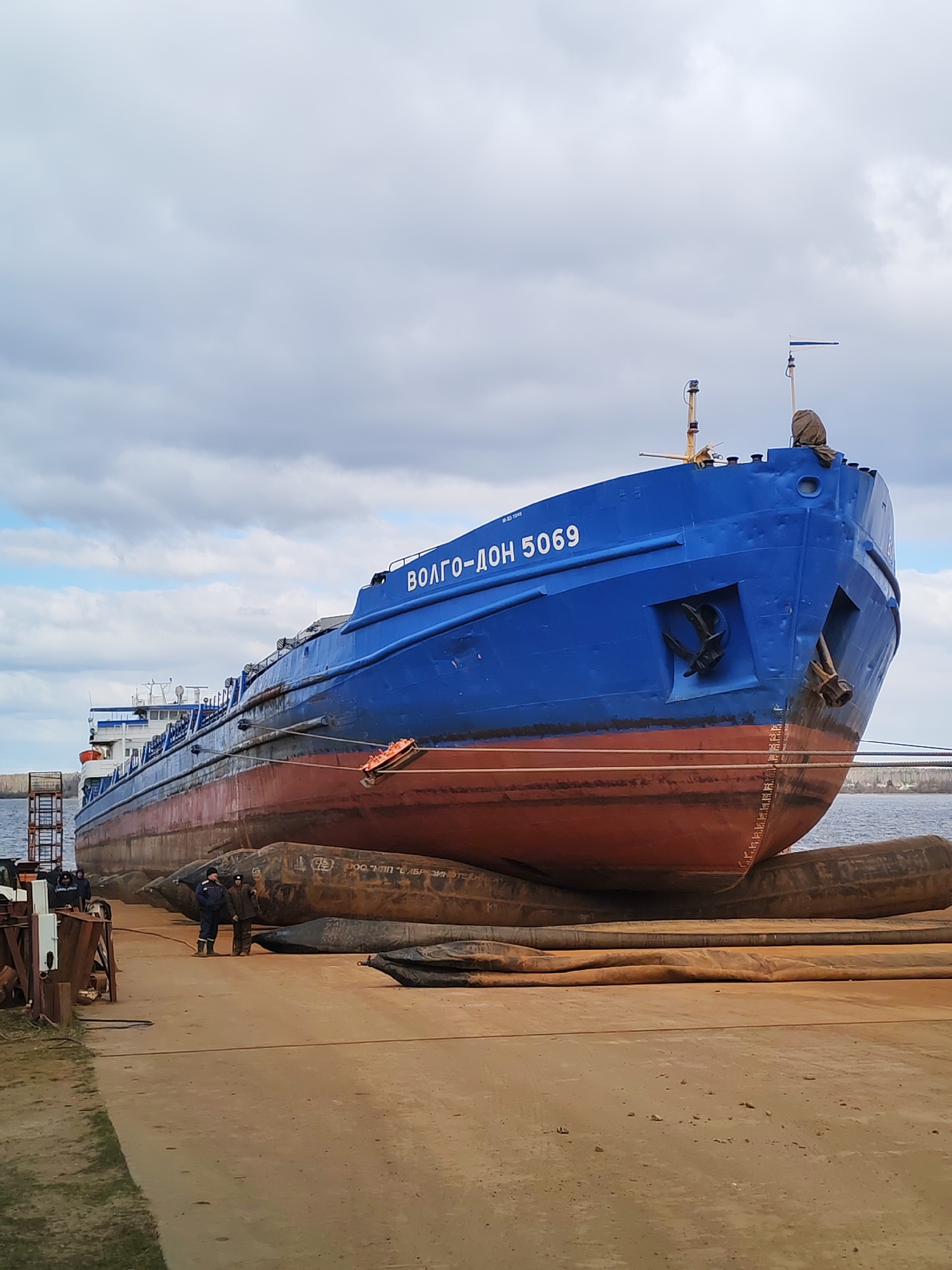 A large, blue and red cargo ship, identified as "ВОЛГО-ДОН 5069" (Volgo-Don 5069) in white Cyrillic text on its bow, is positioned out of the water on what appears to be a dry dock or slipway in Rybinsk, Russia. The vessel's hull, painted a vibrant blue above the waterline and a rust-red below, is supported by numerous large, deflated or partially inflated dark brown/black pneumatic airbags or rollers arranged on the sandy ground. One of these airbags bears the partially visible Cyrillic text "ООО "МПП "СЕВЗИНТЕХ"".
The image captures the ship's front section, featuring prominent anchors on either side of the bow. White draft marks are visible on the red hull near the bow. Several men, dressed in work attire, are present on the ground to the left of the ship, with at least two clearly visible figures standing and observing the operation. A metal scaffolding-like structure is also present on the left side of the frame. In the background, a body of water (likely a river, given the ship's name referencing the Volga-Don system) is visible, leading to a distant forested shoreline. The sky is overcast with gray and white clouds. The scene depicts a daytime industrial operation, likely involving ship maintenance, repair, or movement, under cloudy weather conditions.
