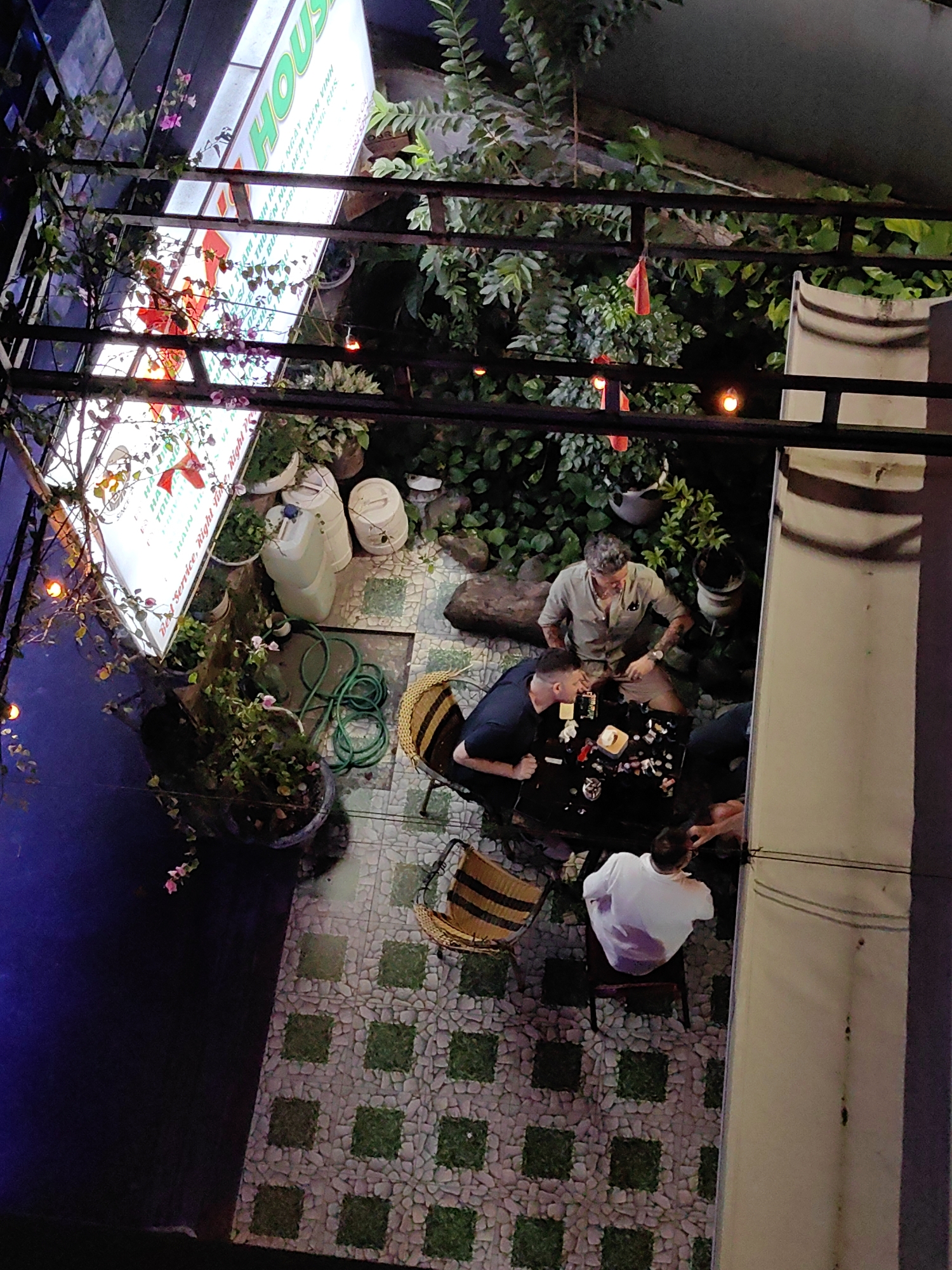 Here's a detailed description of the image:
**General Impression:** The image captures an overhead perspective of a semi-outdoor dining area at night, likely in a commercial setting. The lighting is a mix of artificial illumination and ambient darkness.
**Location:** The presence of the sign indicating "THAIN" and "HOUS" suggests the location is in Vietnam. Given the reference to "Bai Chay Ward," it is likely this scene is located in the Bai Chay Ward area of Vietnam.
**People:** Several individuals are visible around a table.
* One man is wearing a light-colored button-down shirt and shorts.
* Another man is wearing a black shirt.
* A third man is wearing a white t-shirt.
* Other individuals are partially visible around the table.
* The individuals appear to be engaged in conversation or dining.
**Objects:**
* A black table is the central point of the dining area, covered with various objects including bottles, glasses, and a candle.
* Several chairs surround the table, including wicker chairs.
* There are various plants, including potted plants and climbing vegetation.
* Large white containers or tanks are located near the sign.
* A sign is visible, presumably for the business, with the words "HOUS" prominently displayed.
* A green garden hose is visible on the floor.
* Decorative lights and red flags are strung overhead.
* The flooring is made of stones with green patches of grass in between.
**Actions and Interactions:** The focus of the scene is the interaction between the individuals at the table. They are engaged in some form of social activity, possibly eating, drinking, or conversing.
**Environment:** The area has a relaxed, informal atmosphere. The plants and decorative lighting create a pleasant ambiance. The presence of outdoor-style flooring suggests this is an open-air space. Part of the dining area is adjacent to a white building.