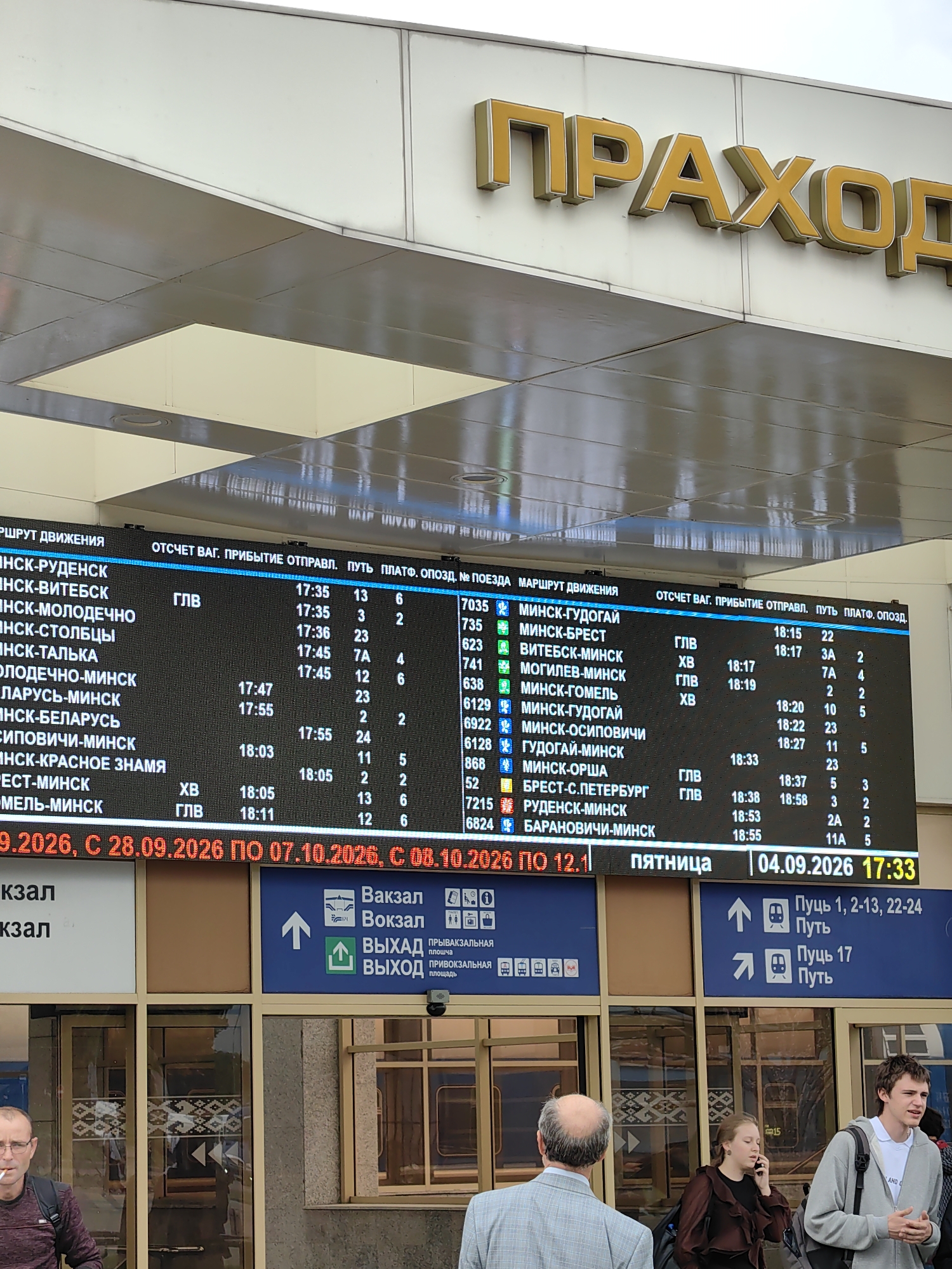 The image shows a street view of a train station in Minsk, Belarus. A large electronic display board lists train departure times and destinations. The board is in Cyrillic script and includes information such as departure times, destination cities, platform numbers, and ticket prices. The display also indicates the current date and time. In the foreground, several people are visible, including a man smoking a cigarette and a woman talking on a mobile phone. The architecture suggests a modern public transportation hub. The overall environment appears to be an outdoor concourse area of the train station.