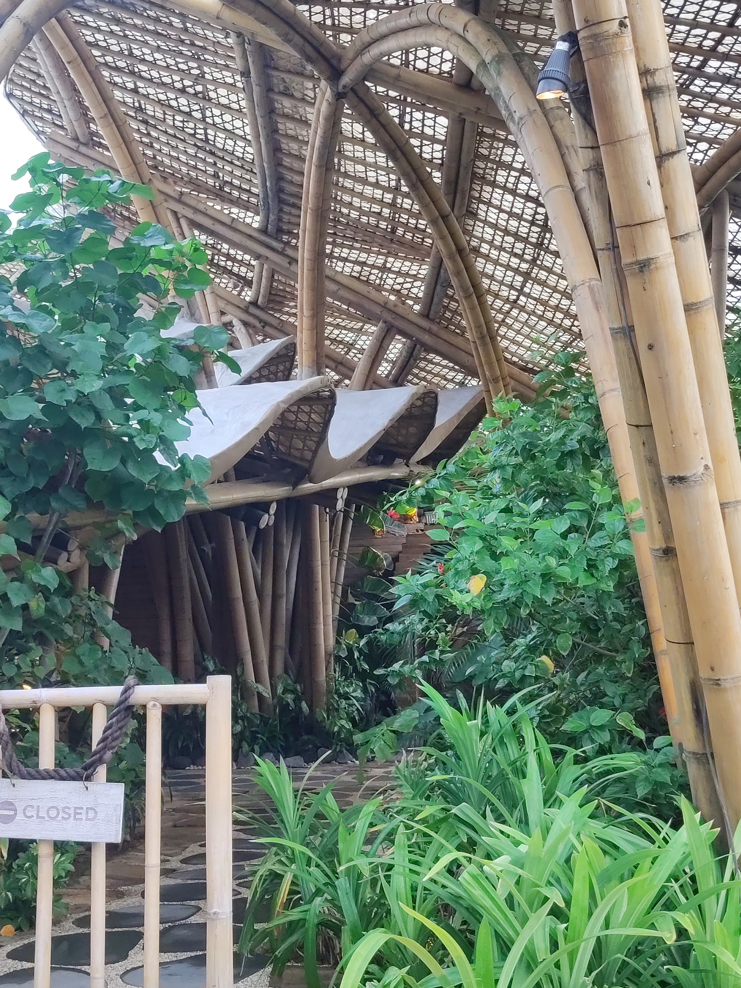 An exterior shot shows a building with a unique bamboo structure. The roof is made of a latticework of bamboo with a translucent covering. Large bamboo poles support the arched roof. Lush green foliage surrounds the entrance, with some red and yellow flowers visible. A pathway made of stone slabs leads towards the building. A white bamboo fence with a "CLOSED" sign hanging from a rope is in the foreground, to the left. The overall impression is of an open-air structure integrated with nature.
