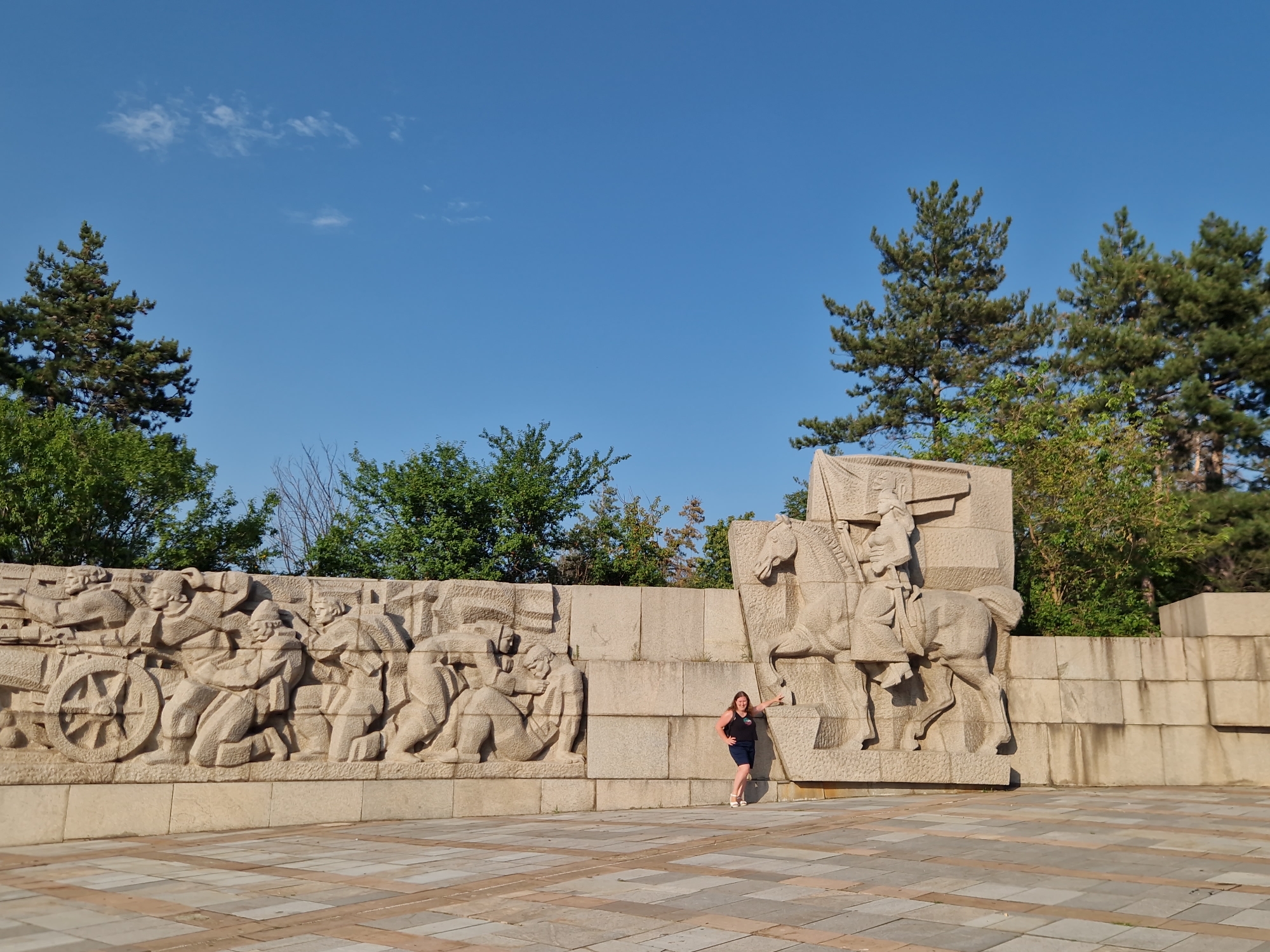 In Panagyurishte, Bulgaria, a woman stands in front of a large, light-colored stone monument featuring bas-relief sculptures. The monument depicts a procession or battle scene with numerous figures and a horse-drawn cart on the left, and a figure on horseback on the right. The woman, wearing a dark sleeveless top and a blue skirt, leans against the monument. The background consists of a clear blue sky with a few wispy clouds and a line of trees, including several tall pine trees. The ground in the foreground is paved with large, square tiles. The monument appears to be of historical significance, likely commemorating an event or figures from Bulgarian history.