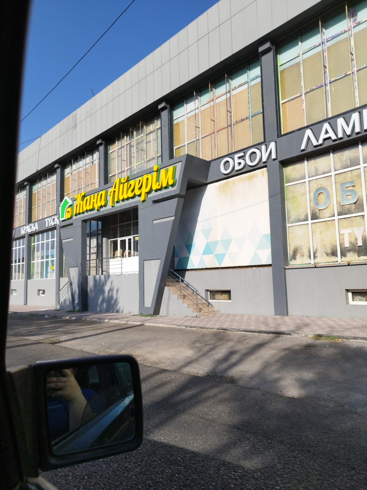 The image shows a building exterior in the city of Shymkent, Kazakhstan. The building has a modern design with large windows and signage. One prominent sign in yellow lettering reads "Жана Шыгыр" which translates to "New Way" or "New Path". Another sign in white lettering reads "ОБОИ", meaning "wallpaper". Further along the building, in large white letters, is "ЛАМИ". On the left side of the building, there are signs that read "КРАСКА" (paint) and "ТУСК" (dull). The building appears to be a retail establishment, possibly a home improvement or decor store. In the foreground, a car's side mirror is visible, reflecting a person's arm and hand. The sky is clear and blue, indicating daylight. The street in front of the building is paved, and shadows from trees are cast on the ground.