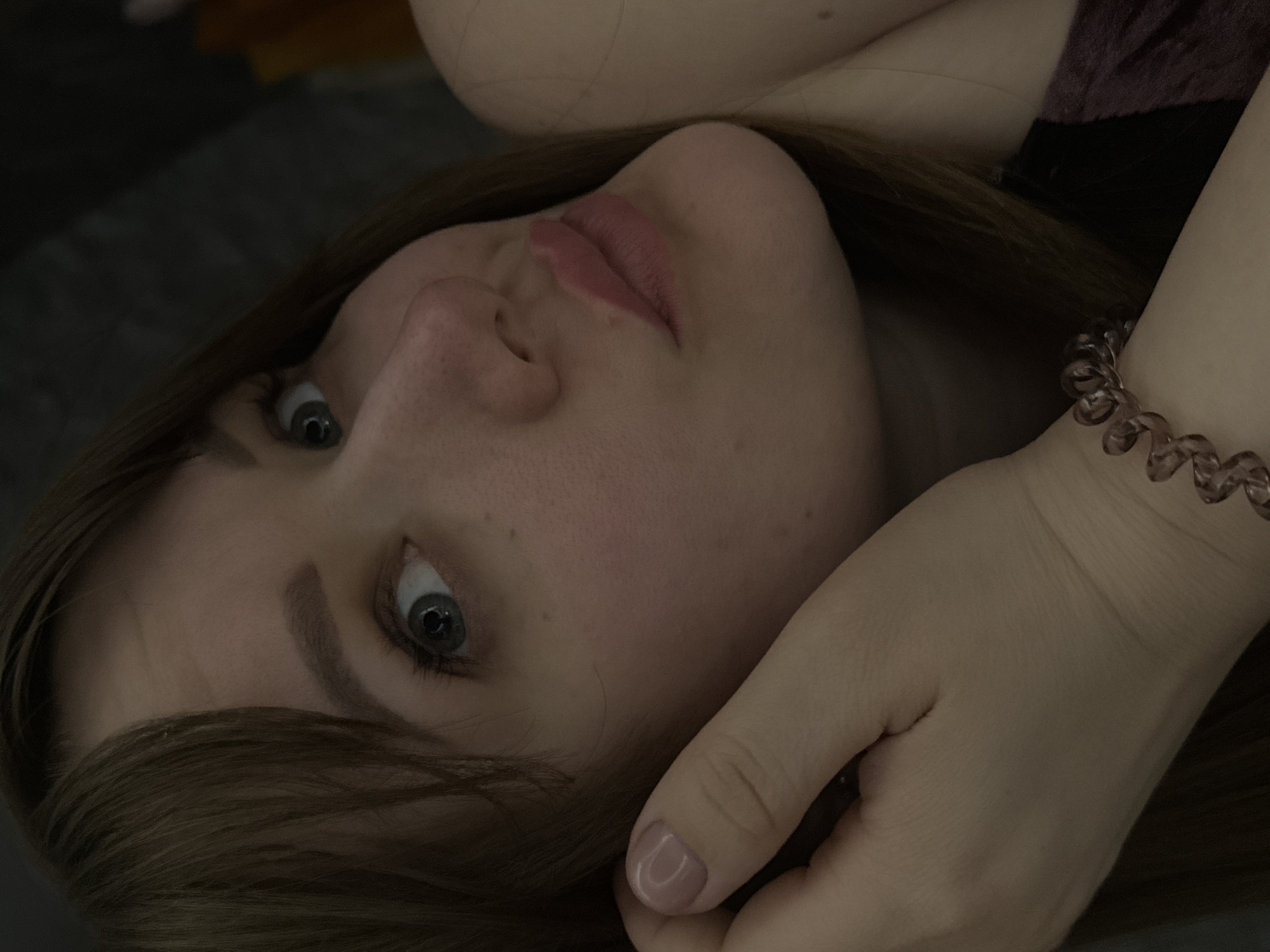 Close-up shot of a woman's face, tilted towards the camera. She has fair skin, blue eyes, and light brown hair. Her lips are closed, with a slight pink hue. A hand is positioned near her face, with a pink spiral hair tie visible around the wrist. The background is dark and out of focus. It is not possible to confirm the location.
