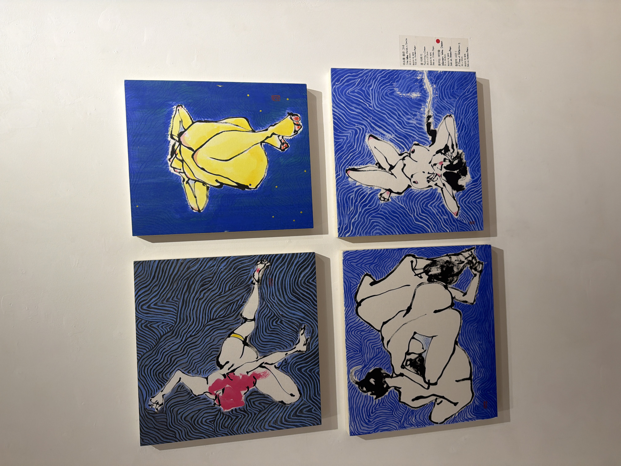 The image shows four paintings displayed on a white wall in what appears to be an art gallery in Seoul, South Korea, based on the text on the exhibition label visible in the lower right quadrant of the image. The paintings are arranged in a two-by-two grid.
The top-left painting features a stylized depiction of a figure with pink hair, rendered in white and black ink, with a yellow accent around the waist. The background is a pattern of wavy blue lines.
The top-right painting shows a figure in yellow, lying on a blue background with small yellow dots, resembling stars. The figure is depicted from the knees up, with limbs crossed.
The bottom-left painting is in black and white, with a black figure partially obscuring a white figure, both rendered in a loose, ink-wash style. The background is the same wavy blue line pattern as the top-left painting.
The bottom-right painting, also in black and white, shows a figure lying upside down with its head angled downwards. The background is the wavy blue line pattern. An exhibition label is affixed to the wall to the right of this painting, providing titles and details in Korean and English, along with a red artist's seal. The overall aesthetic is modern and artistic, with a focus on figurative representation and abstract backgrounds.