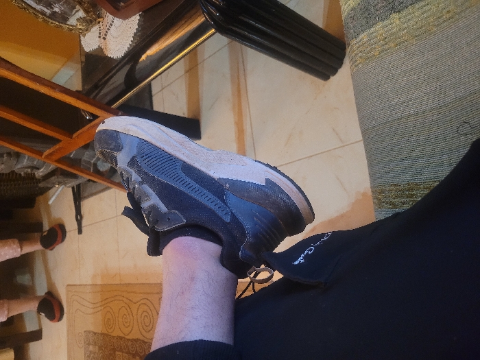 The scene depicts an interior residential setting in the city of Κωστήτσι, Greece. In the immediate foreground, the lower left leg of an individual is positioned, wearing a black and grey athletic shoe with a thick beige-white sole, visibly covered in dirt and dust. The individual's trousers are black, with "Pierre Cardin" embroidered in white script on the upper thigh area. The leg displays visible hair. This individual appears to be seated on a sofa or chair, upholstered in a striped fabric featuring tones of grey, green, and beige.
In the mid-ground, a tiled floor with light beige square tiles and dark grout lines is visible, partially covered by a rectangular rug with a light background and a swirling curvilinear pattern. Further back, another individual's lower legs and feet are present, clad in pink patterned pajama-style trousers and dark red/brown slippers with black soles.
To the right, a dark, reflective table, possibly glass-topped, holds a white crocheted doily and a gold-framed item, likely a picture frame. Next to the table, a wooden, ladder-like structure, possibly a chair or decorative object, stands. A dark, cylindrical object rests on the floor beside the table. The background features a solid yellow-ochre painted wall. The overall impression is that of a casually occupied living space.
