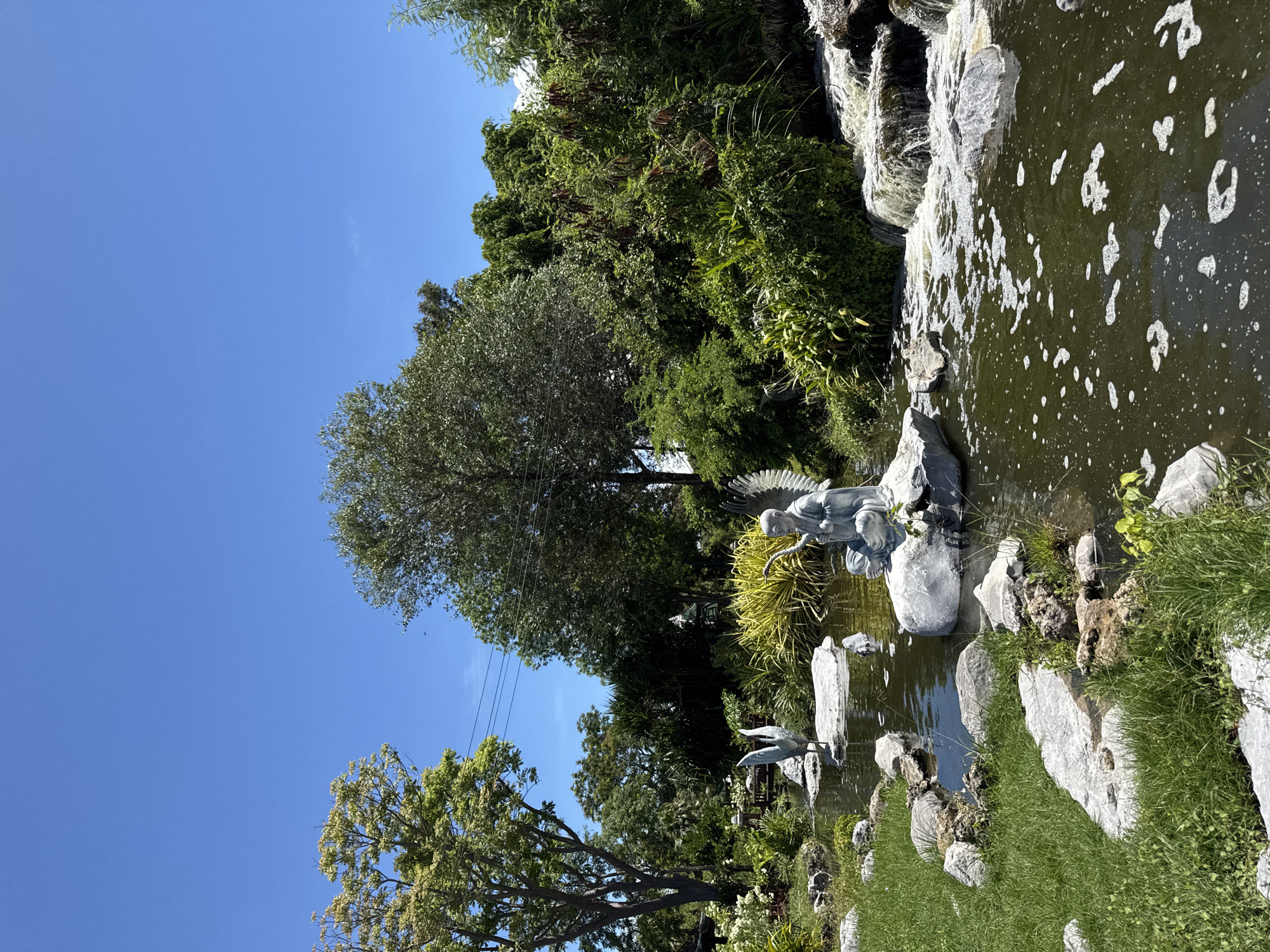 The image depicts a garden scene in Bang Pu Subdistrict Municipality, Thailand. A pond with an artificial waterfall borders lush greenery and trees under a clear blue sky. Stone arrangements surround the pond, which also contains two sculptures: one of an angel and another of a heron. Water flows in the pond. There are also power lines.