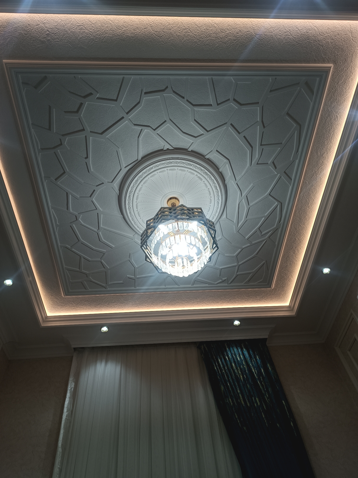 A detailed, forensic, matter-of-fact description of the scene is as follows:
The image shows an interior shot of a ceiling with a chandelier. The ceiling is decorated with a geometric pattern and recessed lighting around its perimeter. The chandelier is ornate, with multiple tiers of crystal-like elements and a metallic frame. Below the ceiling, partially visible, are sheer white curtains and darker, textured curtains. The wall appears to be textured, with a subtle pattern. The scene is likely a residential or commercial interior space. There are no people or overt actions depicted. The lighting in the room originates from the recessed ceiling lights and the chandelier, creating a warm and ambient atmosphere. The photograph is taken from a low angle, looking upwards towards the ceiling. The location is identified as Myrza-Ake, Kyrgyzstan.