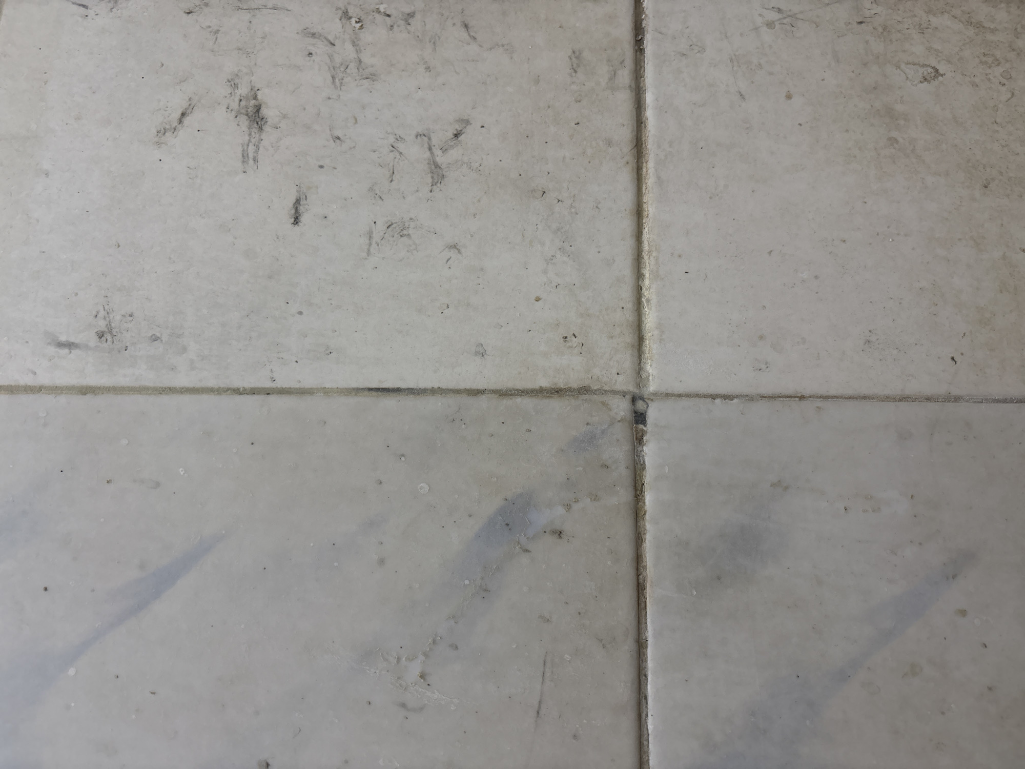 The image shows a close-up, overhead view of a dirty tiled floor. There are four square tiles visible, meeting in the center of the frame. The tiles are a light, neutral color, possibly white or a very pale gray. The surface of the tiles is smudged and stained with dark, irregular marks, appearing to be dirt, scuff marks, or possibly footprints. Some tiles have streaks of lighter gray discoloration, potentially from water marks or dried spills. The grout lines between the tiles are visible and appear slightly darker and more worn than the tiles themselves. The overall impression is one of neglect or heavy use, indicating the floor has not been recently cleaned. There are no people, animals, or significant objects visible in the frame, focusing solely on the texture and condition of the floor. This image does not provide any information that can be used to determine its location in Palimanan, Indonesia, or any other specific geographical context.