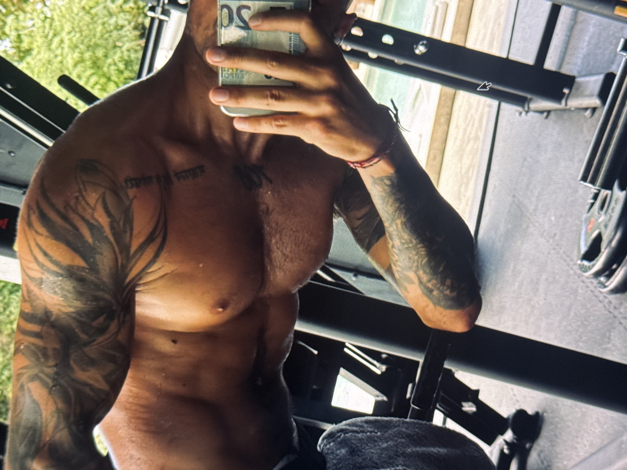 The image captures a close-up view of a muscular individual's torso and arm, heavily tattooed with tribal or abstract patterns. The skin appears to be sweating, suggesting recent physical exertion, possibly in a gym setting. The person is holding a phone in their right hand, which is partially obscured by their fingers. A small, thin bracelet with red and black threads is visible on their left wrist. The background is blurred, but hints of green foliage and a swimming pool suggest an outdoor or semi-outdoor environment, possibly a resort or a high-end gym in a tropical location. The lighting is natural, coming from the side, casting shadows that accentuate the musculature. The overall impression is one of fitness and a relaxed, possibly vacation-like atmosphere. The location is identified as Kuta Selatan, Indonesia, known for its beaches and resorts.