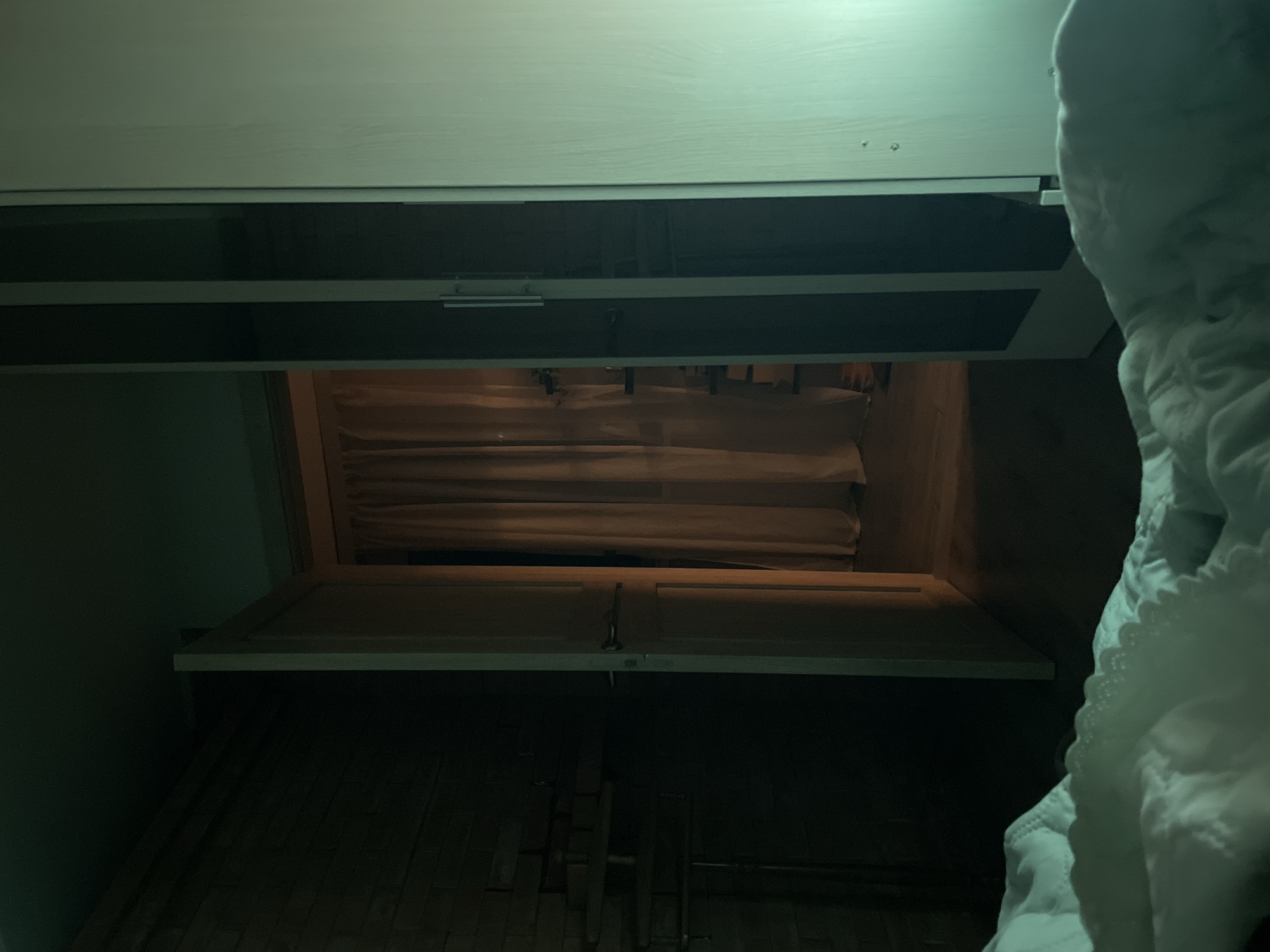 The image depicts the interior of an attic space in Sestroretsk, Russia, as viewed from a low angle. The scene is dimly lit with a greenish hue. A partially opened attic access panel is visible, framed by two white painted surfaces. A section of folded brown insulation material is visible inside the attic space. Below the access panel, what appears to be unfinished wood planks can be seen. The bottom right corner of the image features a white, heavily textured, possibly quilted material.
