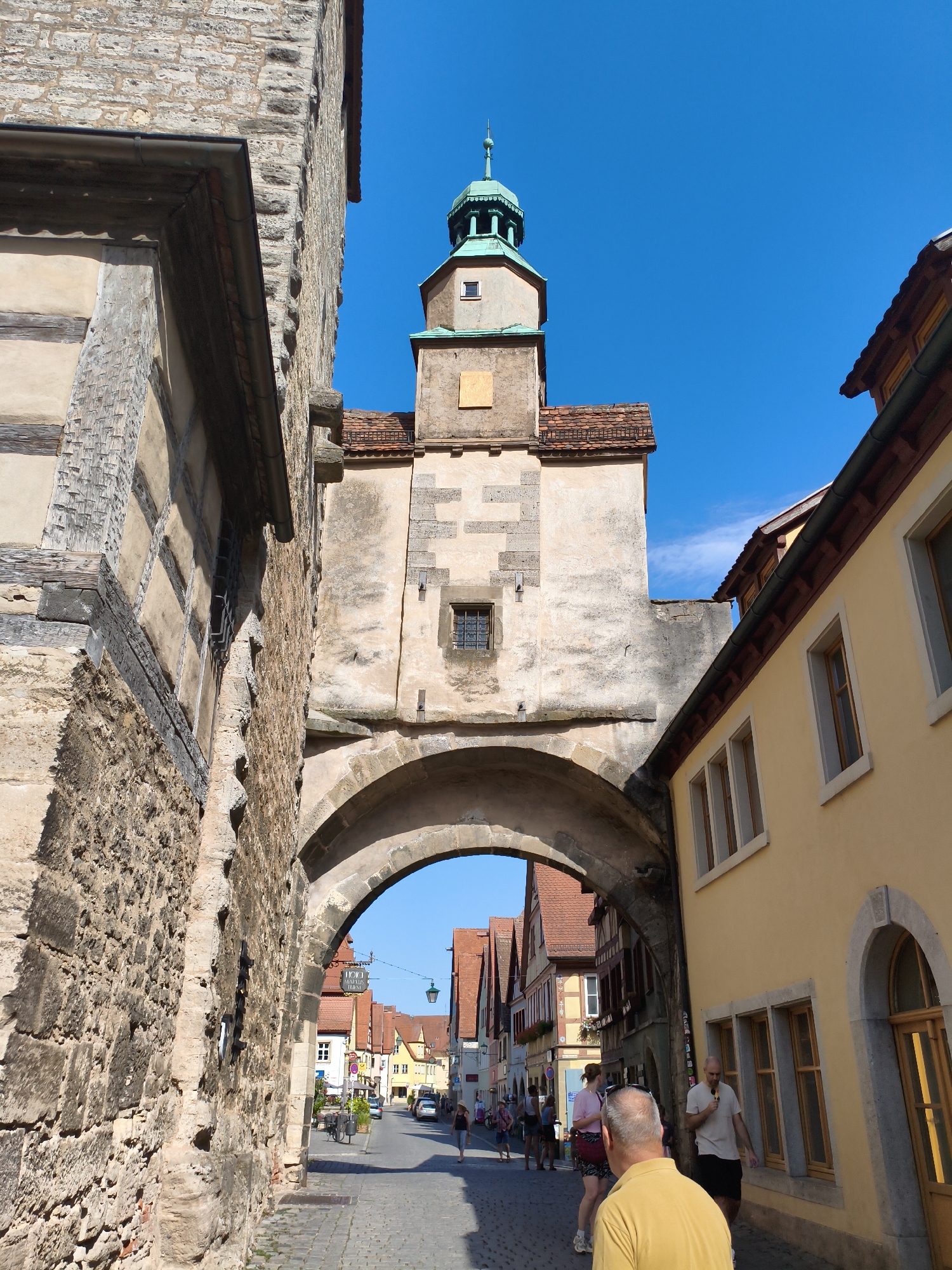 The image captures a view through an archway in Rothenburg ob der Tauber, Germany. The archway, constructed of stone, leads to a cobblestone street lined with historic buildings. The architecture is typical of a medieval German town, with half-timbered structures and red-tiled roofs. A prominent tower with a green spire rises in the background, characteristic of the city's well-preserved medieval center.
Several people are visible on the street. In the foreground, a man in a yellow shirt is facing away from the camera. To his left, two women are walking together. Further down the street, more individuals can be seen, some walking and others possibly shopping. Vehicles, including a bicycle and cars, are also present on the street.
The sky is clear and blue, indicating fair weather. The sunlight casts shadows, highlighting the textures of the stone buildings and the cobblestone pavement. The overall scene conveys a sense of a bustling tourist destination, showcasing the charm and historical significance of Rothenburg ob der Tauber.
