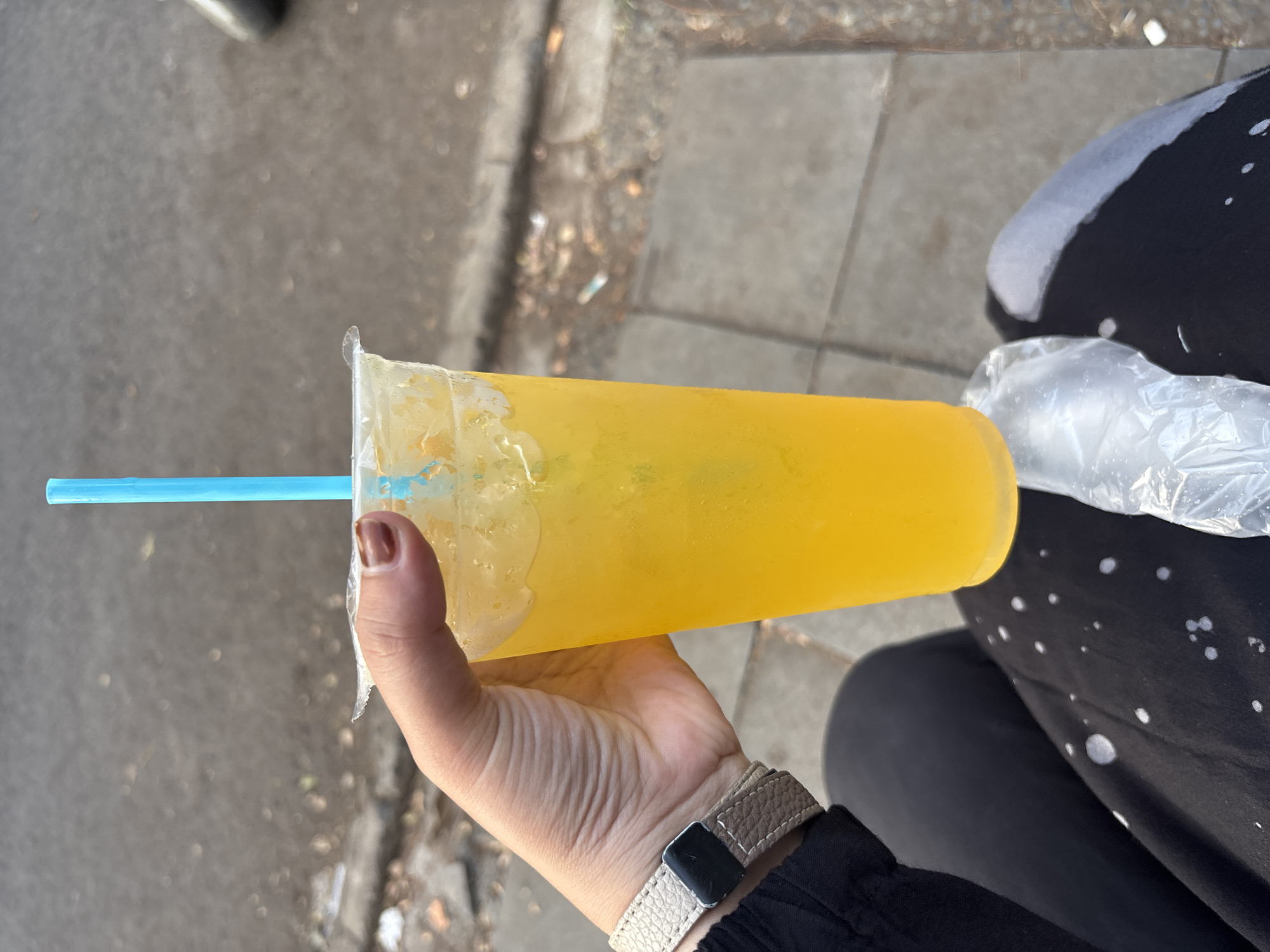 A person's hand, with brown nail polish, holds a clear plastic cup filled with a bright orange beverage and ice. A blue straw extends from the cup. The person is wearing a black long-sleeved shirt and a watch with a beige band. They are sitting down, with their legs crossed, wearing black pants with white circular patterns. A clear plastic bag is visible near their lap. The background suggests an outdoor urban setting with a grey paved surface and some debris. This scene could be located on a street or sidewalk in Cirebon, Indonesia.