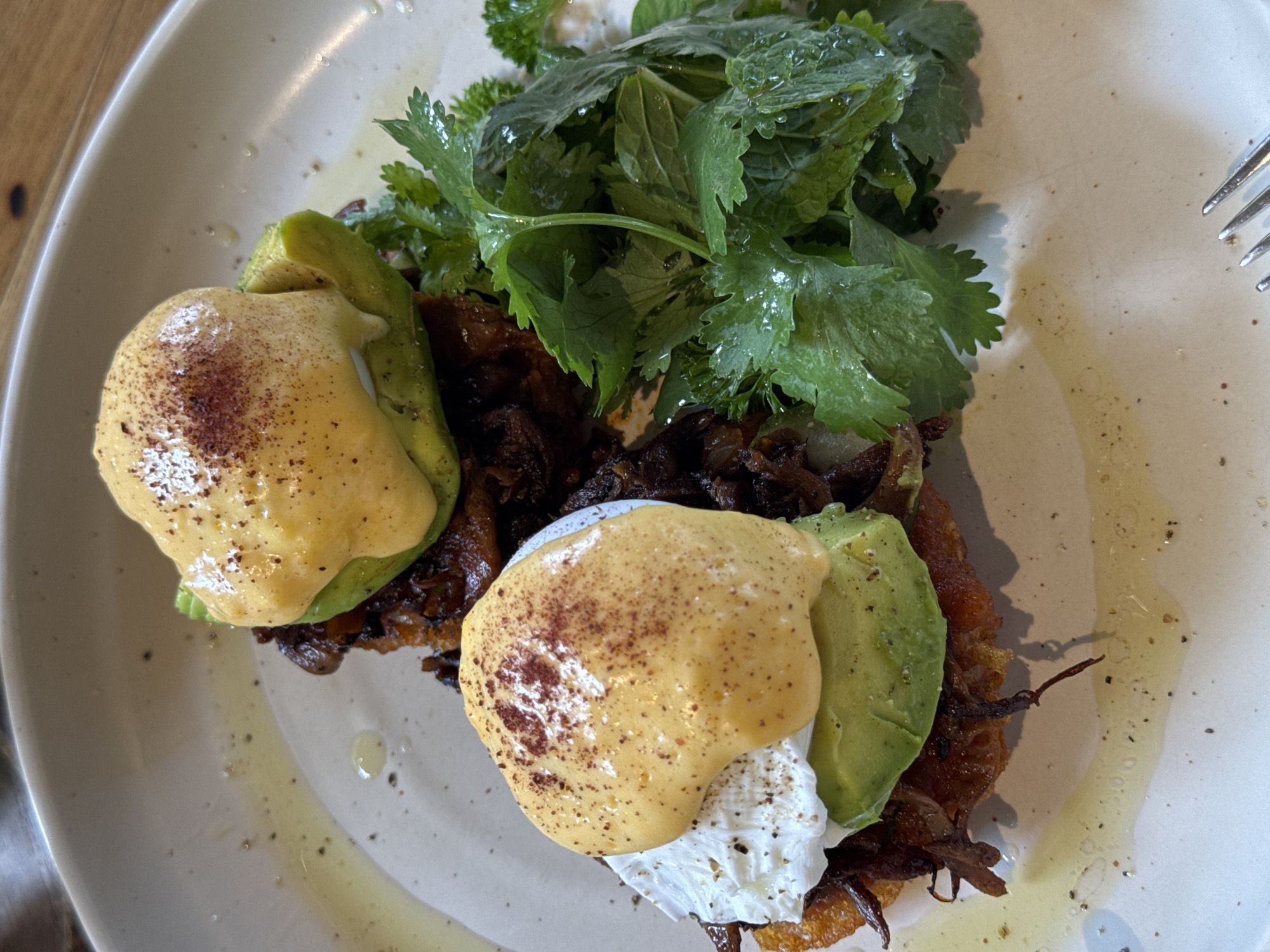 A close-up shot showcases a plate of food, featuring two portions of what appears to be a breakfast dish. Each portion consists of a base, possibly a potato or hash brown, topped with sliced avocado, a poached egg, and a generous drizzle of hollandaise sauce. The dish is garnished with a sprinkle of paprika or chili powder. To the right of the food, a bunch of fresh green herbs, likely cilantro, adds a vibrant contrast. The plate itself is off-white with a subtle speckled pattern, and a small amount of oil and black pepper are visible on its surface. The lighting suggests an indoor setting, possibly a restaurant, with a hint of a wooden surface in the background. There is no indication of the city of Peleana, Sri Lanka within the image.