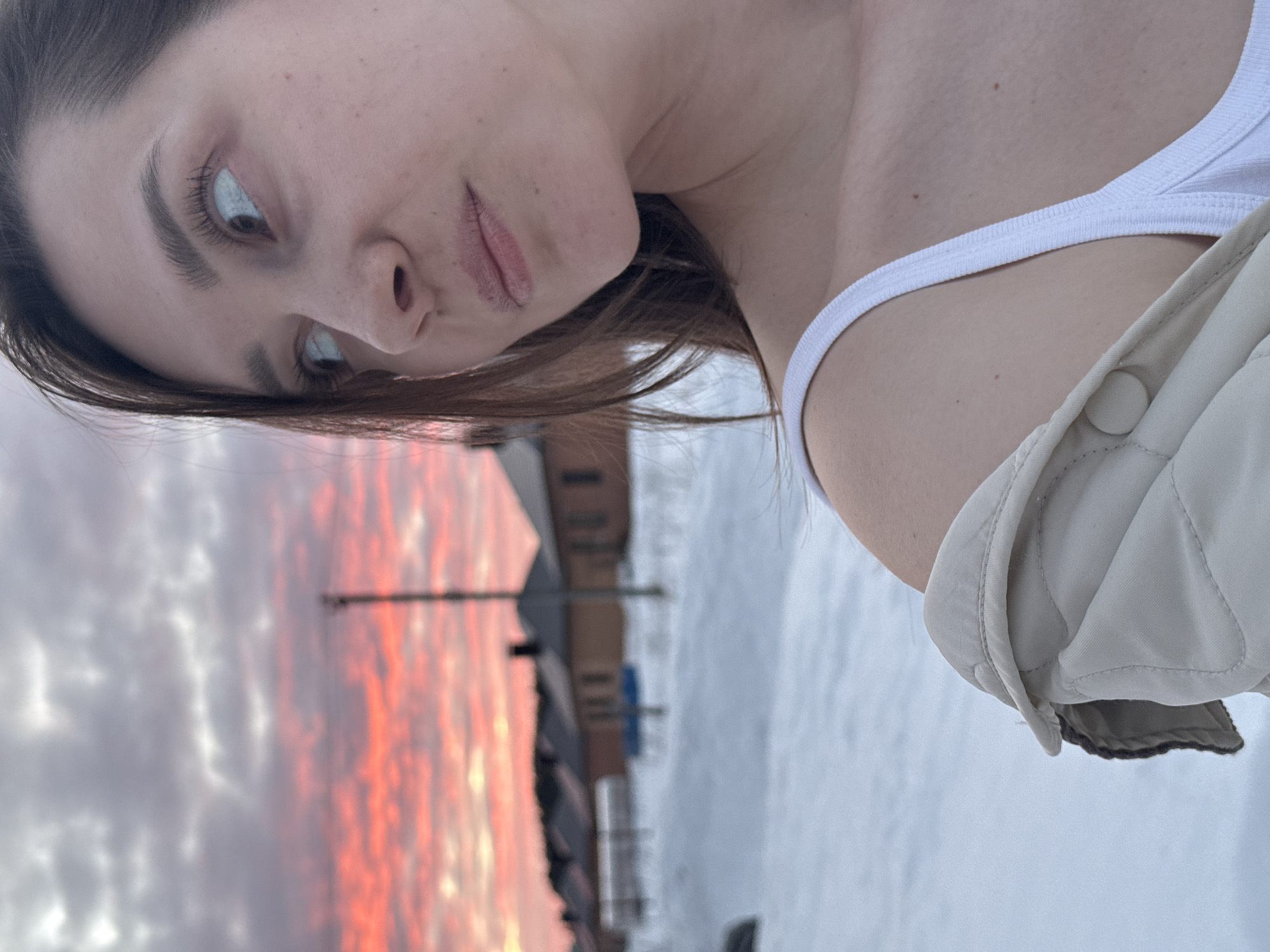 The image depicts a female individual in a snowy residential area of Chaikovsky, Russia, during what appears to be either sunrise or sunset. The subject is positioned in the foreground and occupies the right half of the frame, showcasing her fair skin, dark brown hair, and visible freckles on her cheek. Her right eye is distinctly visible, directed upward and to the left with a contemplative gaze. She wears a white ribbed tank top, partially obscured by a light beige quilted jacket draped over her left shoulder. In the midground, a row of single-story houses with dark roofs and light-colored facades is visible against the snow-covered ground. A slender, dark utility pole stands vertically to the left of the houses. The sky in the background displays vibrant orange and red cloud formations near the horizon, transitioning to lighter grey clouds higher in the frame, indicating a low sun angle.