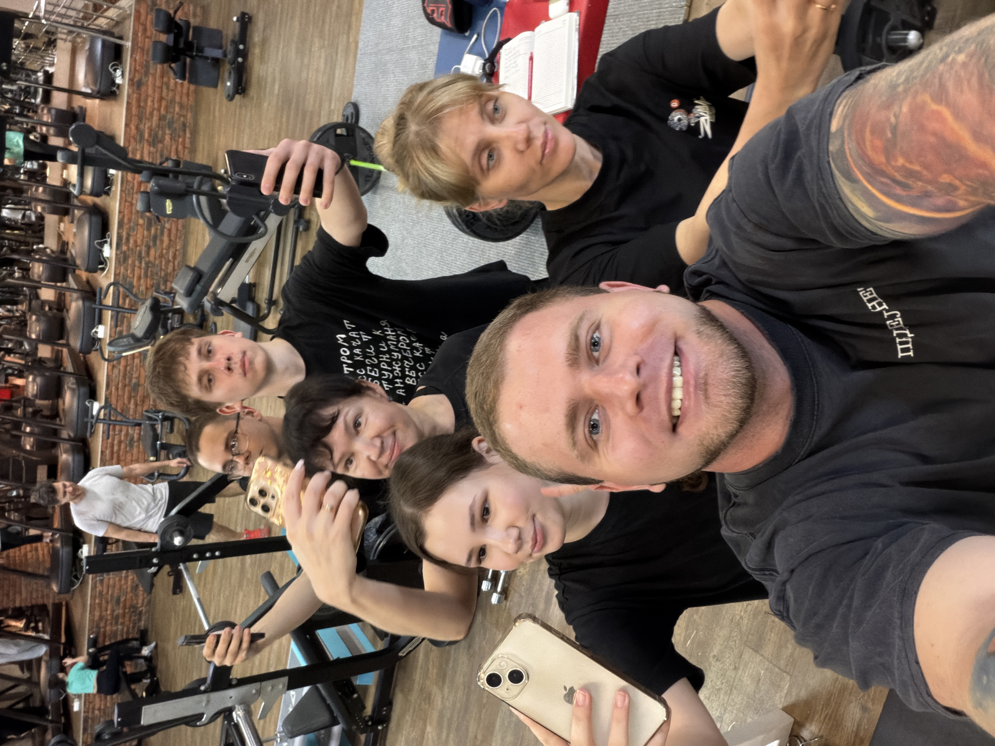 A group of six individuals poses for a selfie inside what appears to be a gym. The picture is taken at a skewed angle, showing the individuals clustered together. All six individuals are wearing dark clothing, and at least three of them are holding smartphones. The gym setting is evident by the presence of exercise equipment visible in the background. From signs in the photo, the location of the gym appears to be Chaikovsky, Russia. Other individuals are present in the background of the image, also seemingly using the gym equipment. The floor has a wooden laminate finish. A red brick wall is also visible.