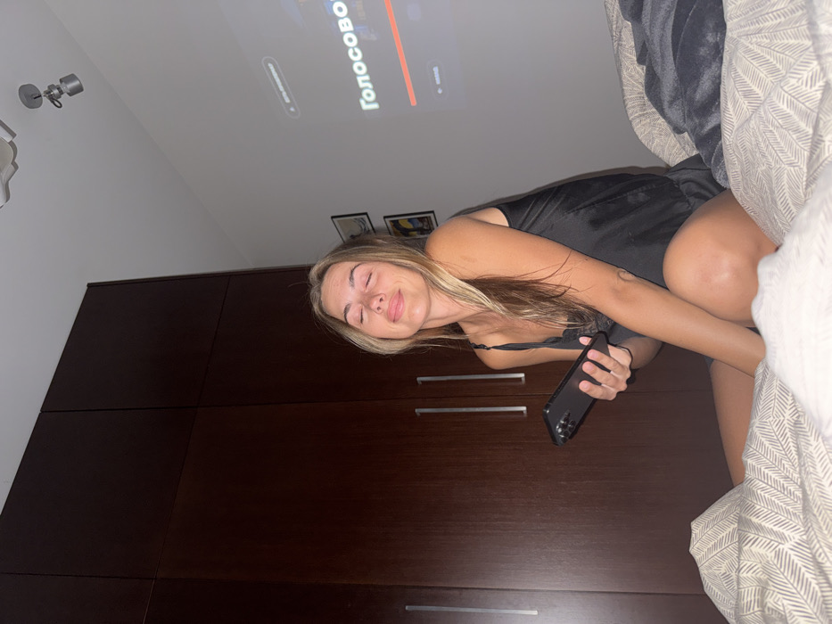 A fair-skinned young woman with long blonde hair reclines on a bed, holding a smartphone. She wears a black silky top. Behind her, a tall dark wood cabinet dominates the scene. On the ceiling, a light fixture and the faint projection of a word in Cyrillic script are visible. The bed is covered with textured bedding in neutral tones.